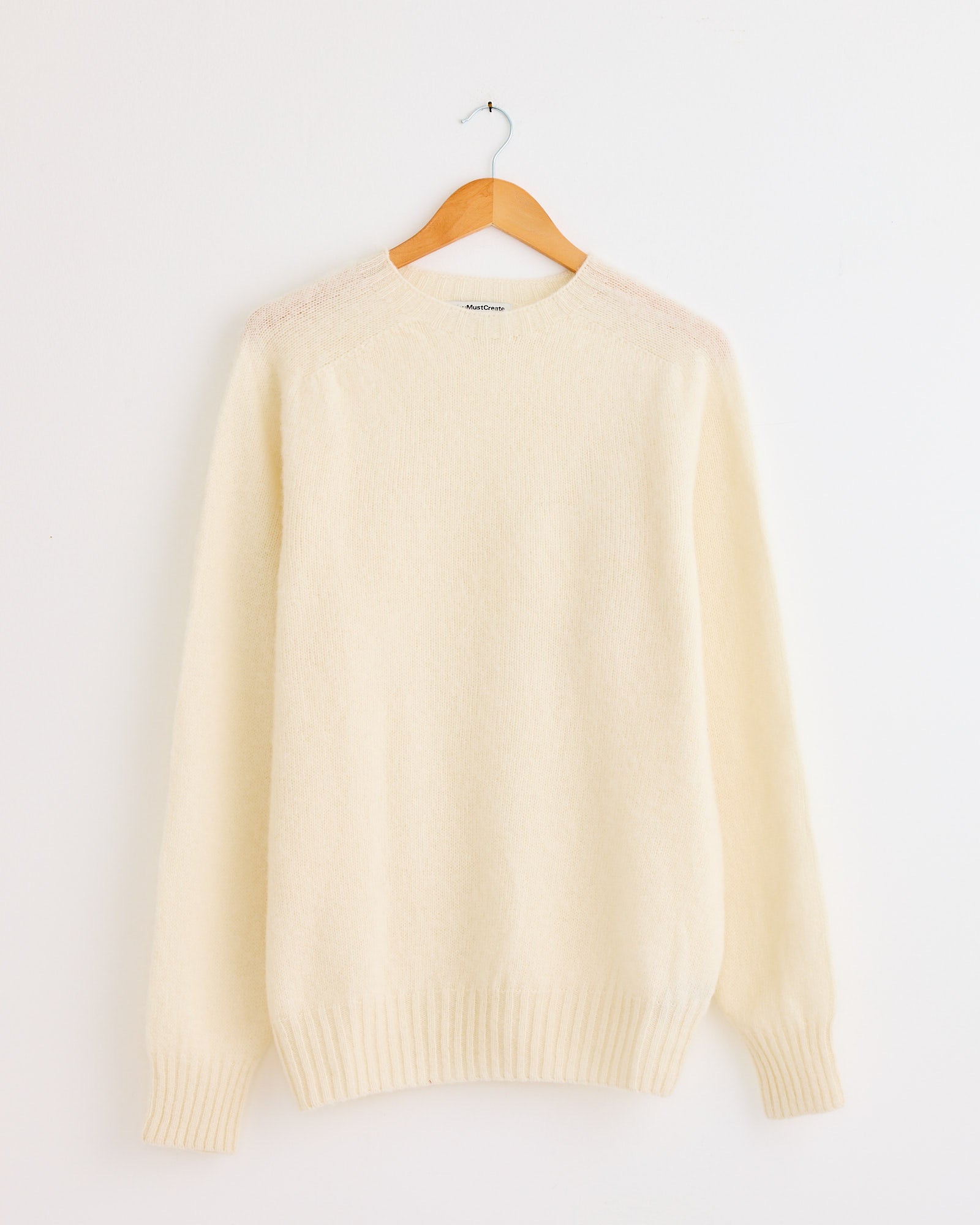The YMC Suedehead Knit in Cream, a long-sleeve sweater, hangs on a wooden hanger against a plain white wall. For fit or sizing questions, our online chat is ready to help.