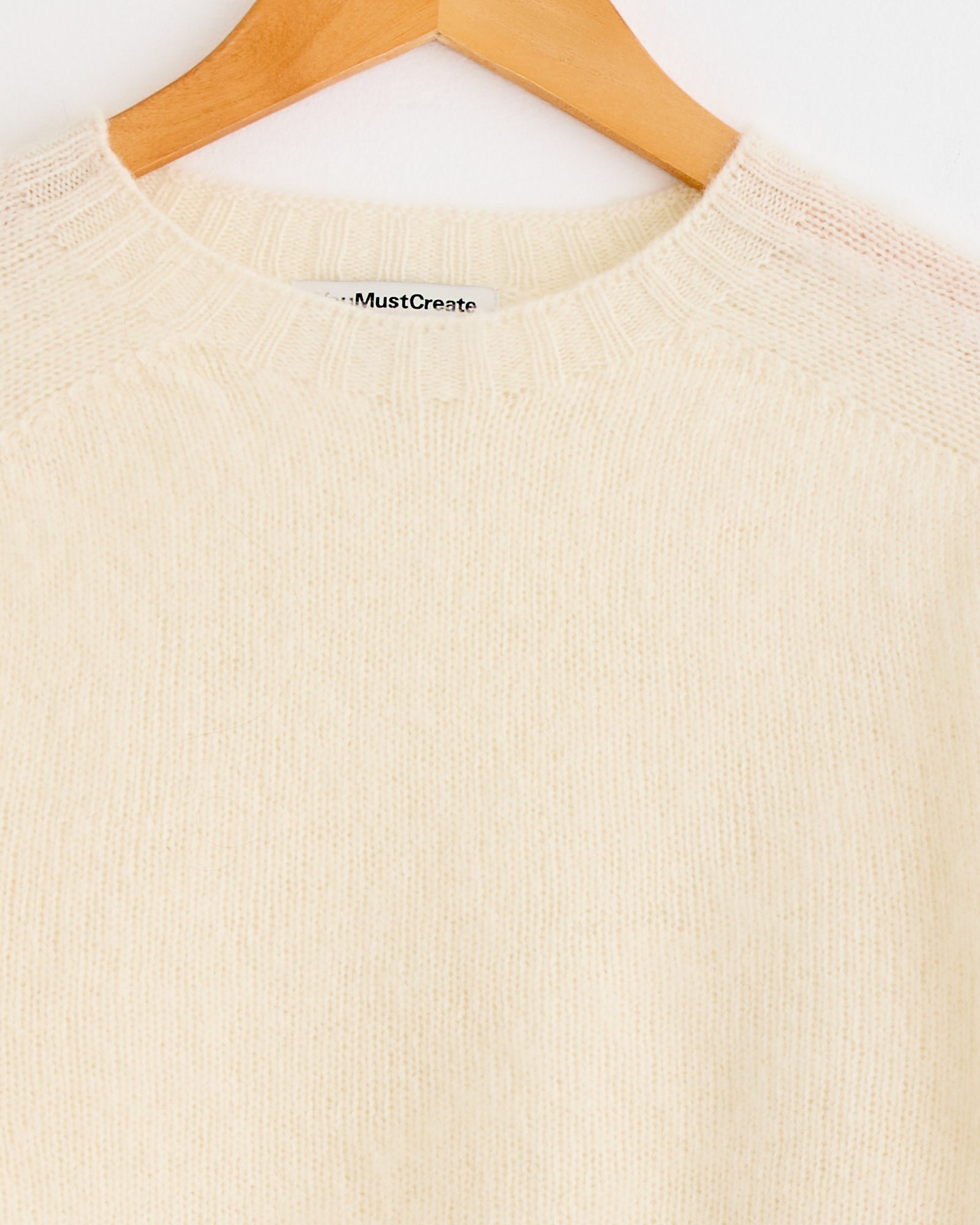 A close-up of the YMC Suedehead Knit in Cream on a wooden hanger, featuring the upper chest, neckline, shoulders, and textured knit. A small label is inside the collar. For sizing or fit questions, please use our online chat.
