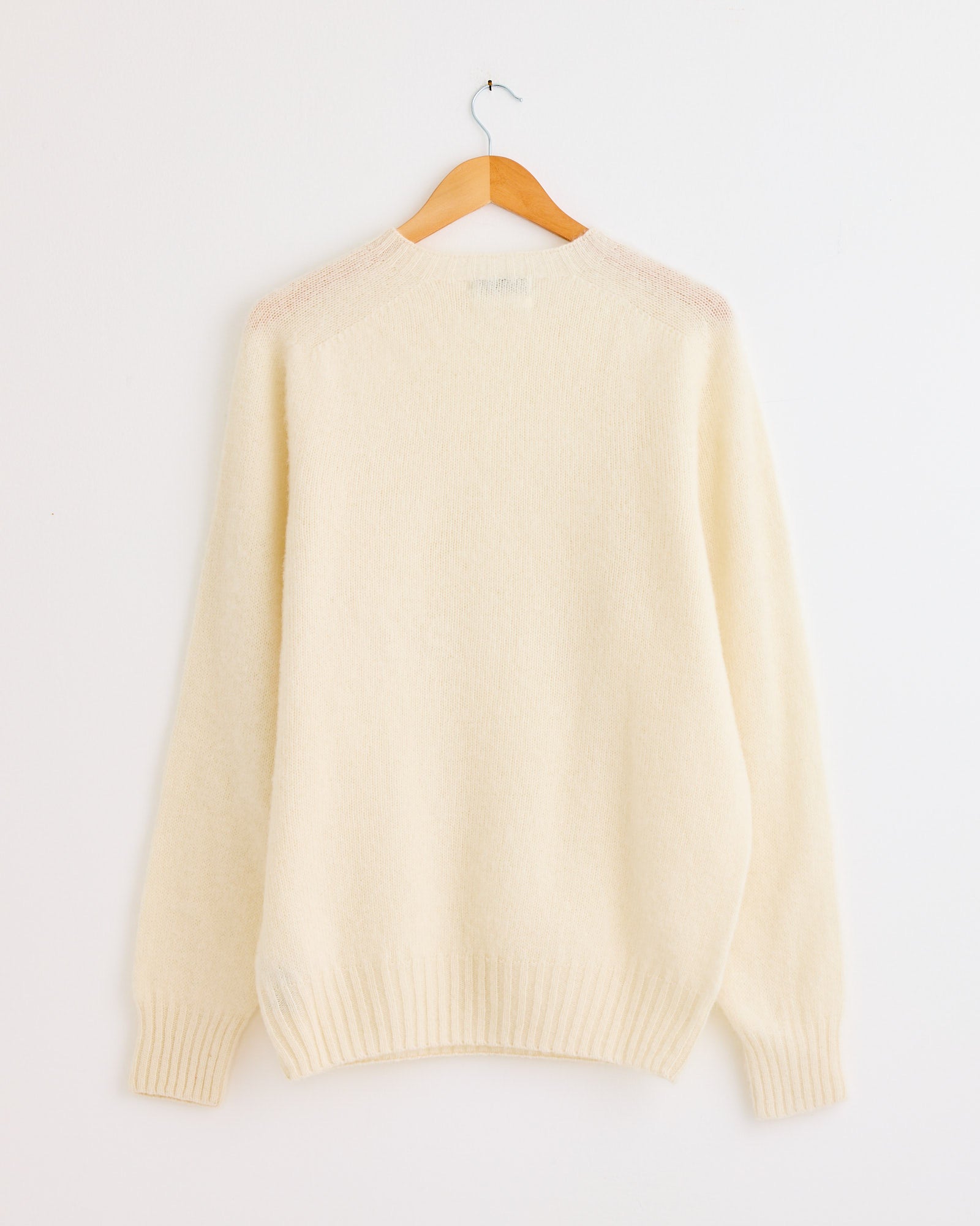 The Suedehead Knit in Cream by YMC, a long-sleeve wool sweater with ribbed cuffs and hem, hangs on a wooden hanger against a white wall. For fit or sizing help, contact us via online chat.