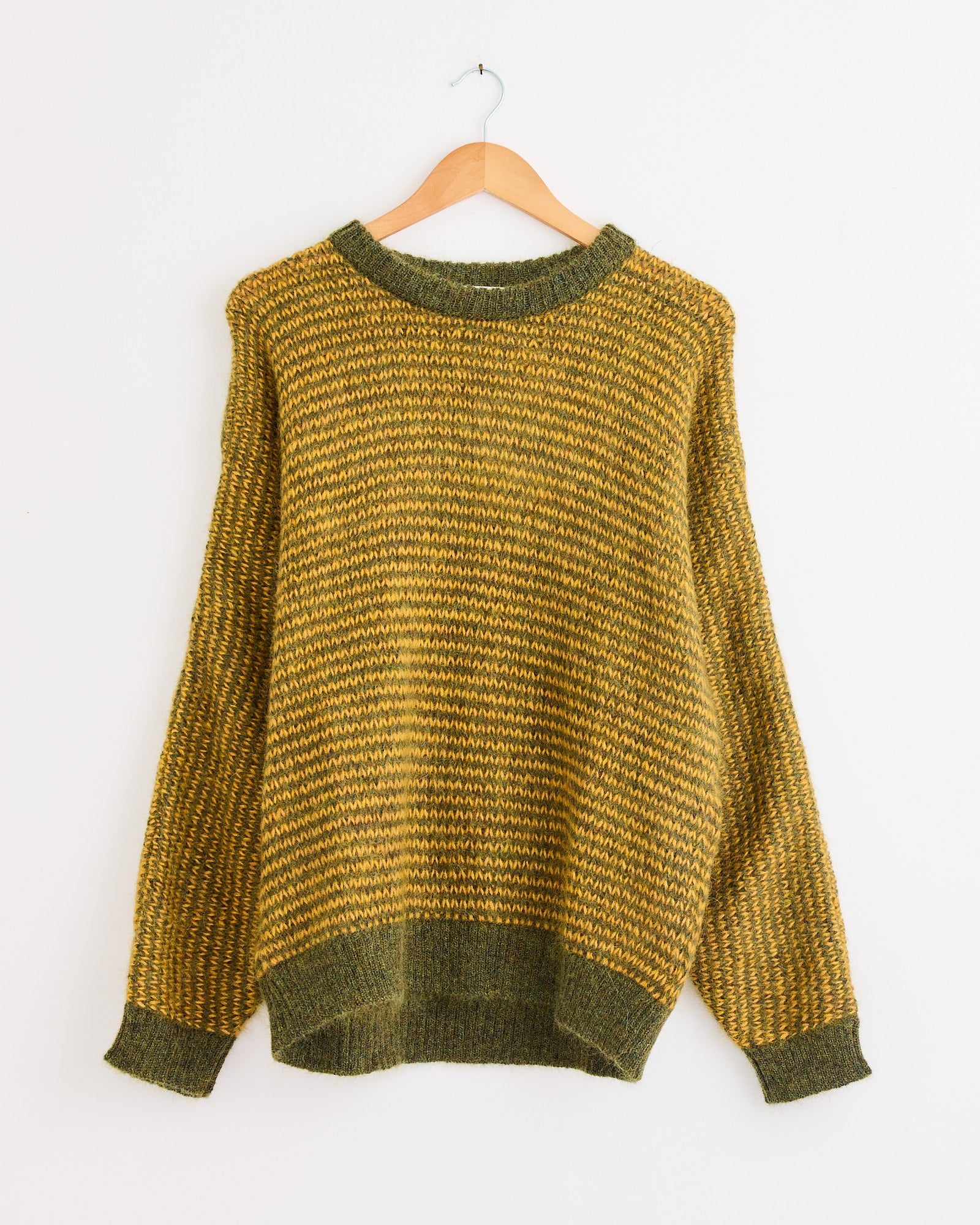 The YMC Black Palm Knit in Yellow/Green, featuring long sleeves and a crew neckline, hangs on a wooden hanger against a white wall. For sizing or fit questions, our online chat is here to help.