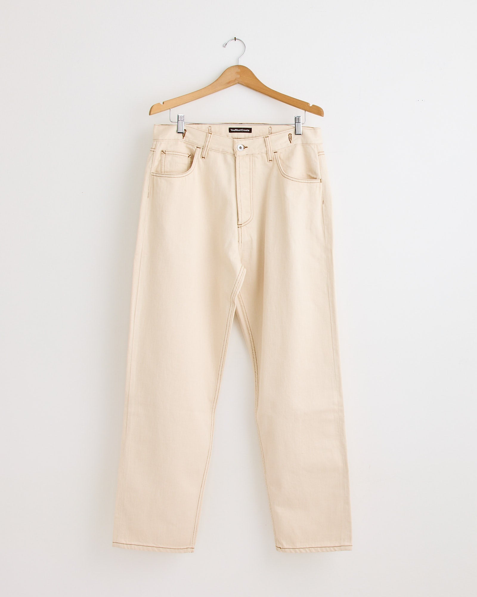 The YMC Bez Jean in Ecru, a high-waisted, straight-leg pair in light beige, hangs on a wooden hanger against a plain white wall—perfect for any fit.