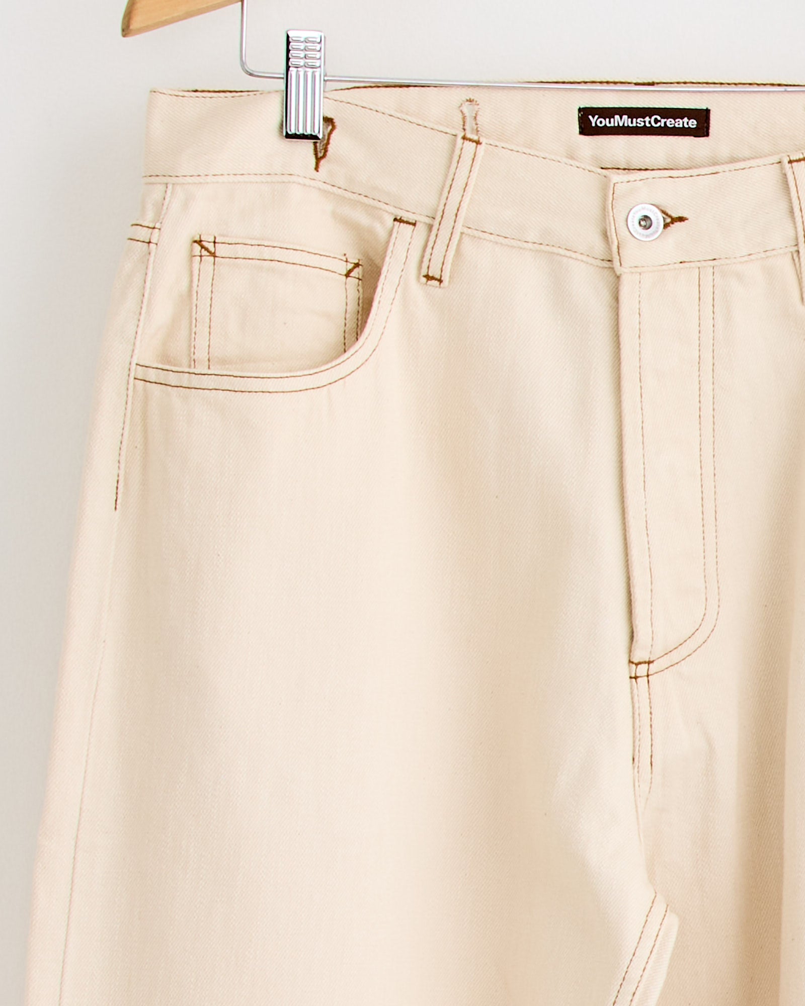 Close-up of the Bez Jean in Ecru by YMC, showing brown stitching, a front pocket, and an inner waistband label. Contact online chat for sizing and fit details.