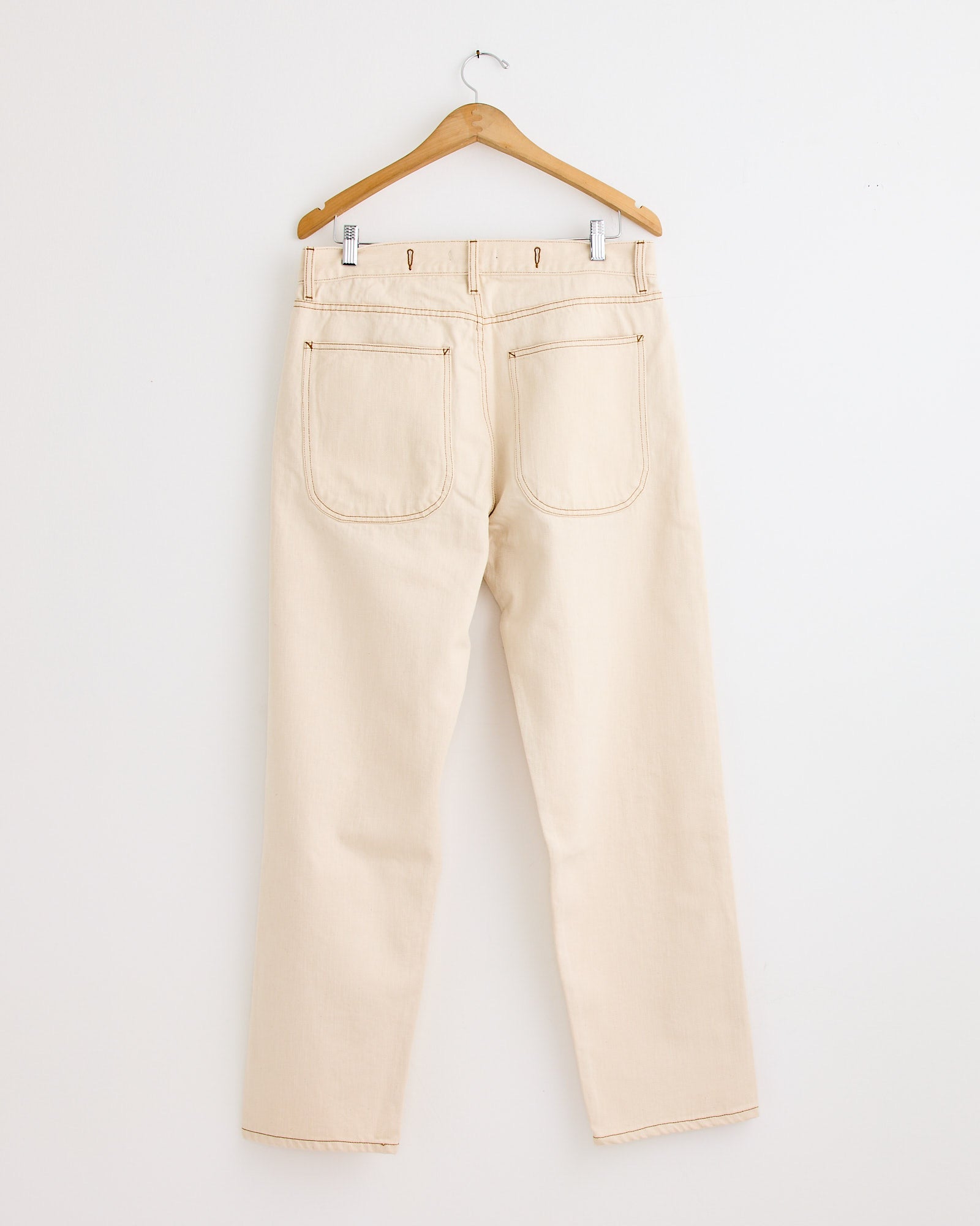 The YMC Bez Jean in Ecru features straight legs and large back pockets, hanging on a wooden hanger against a white wall—ideal for those who value timeless style and effortless sizing.