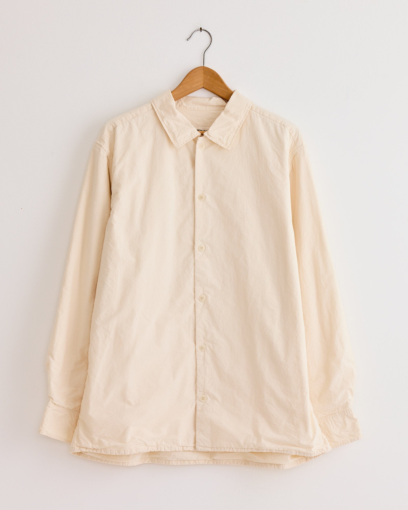 The Casey Casey Mili Shirt in Natural, a long-sleeved style crafted from high density paper cotton, hangs on a wooden hanger against a plain white wall.