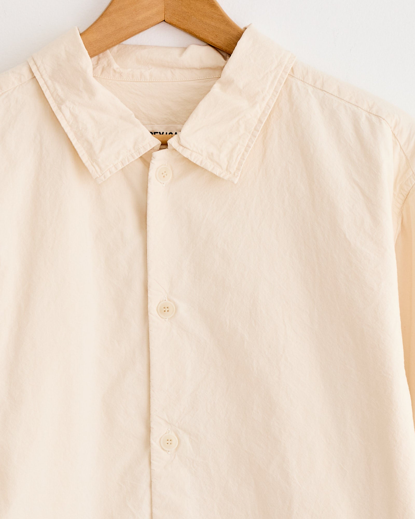 A close-up of the Casey Casey Mili Shirt in Natural, a relaxed fit style made from high density paper cotton with a collar and three visible buttons, shown on a wooden hanger against a white backdrop.