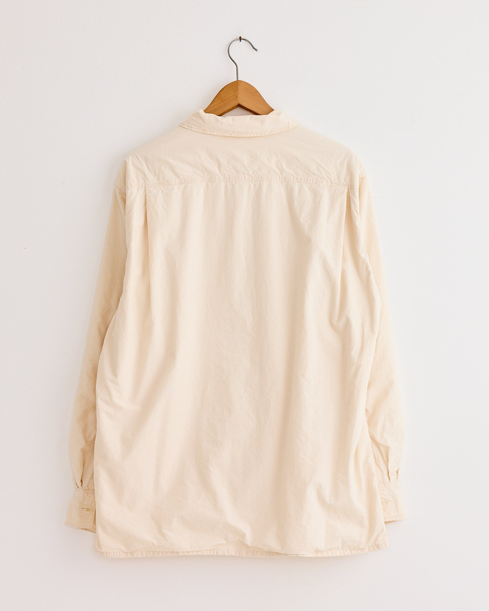 The Casey Casey Mili Shirt in Natural, crafted from high-density paper cotton with long sleeves, hangs on a wooden hanger against a plain white wall and is shown from the back.