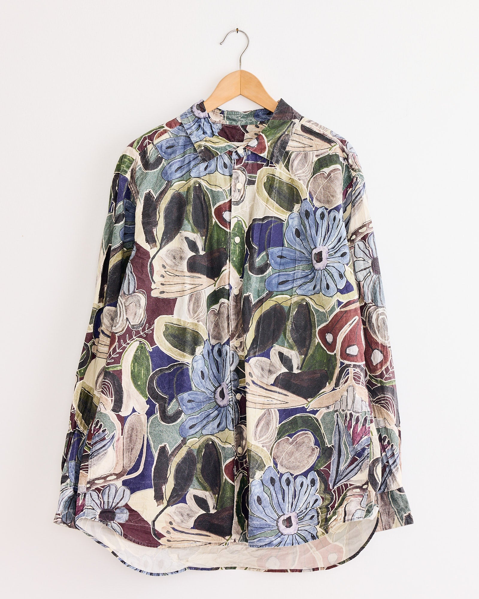 The Big Raccourcie Shirt in Brown by Casey Casey, a long-sleeve button-up, hangs on a wooden hanger against a white wall.