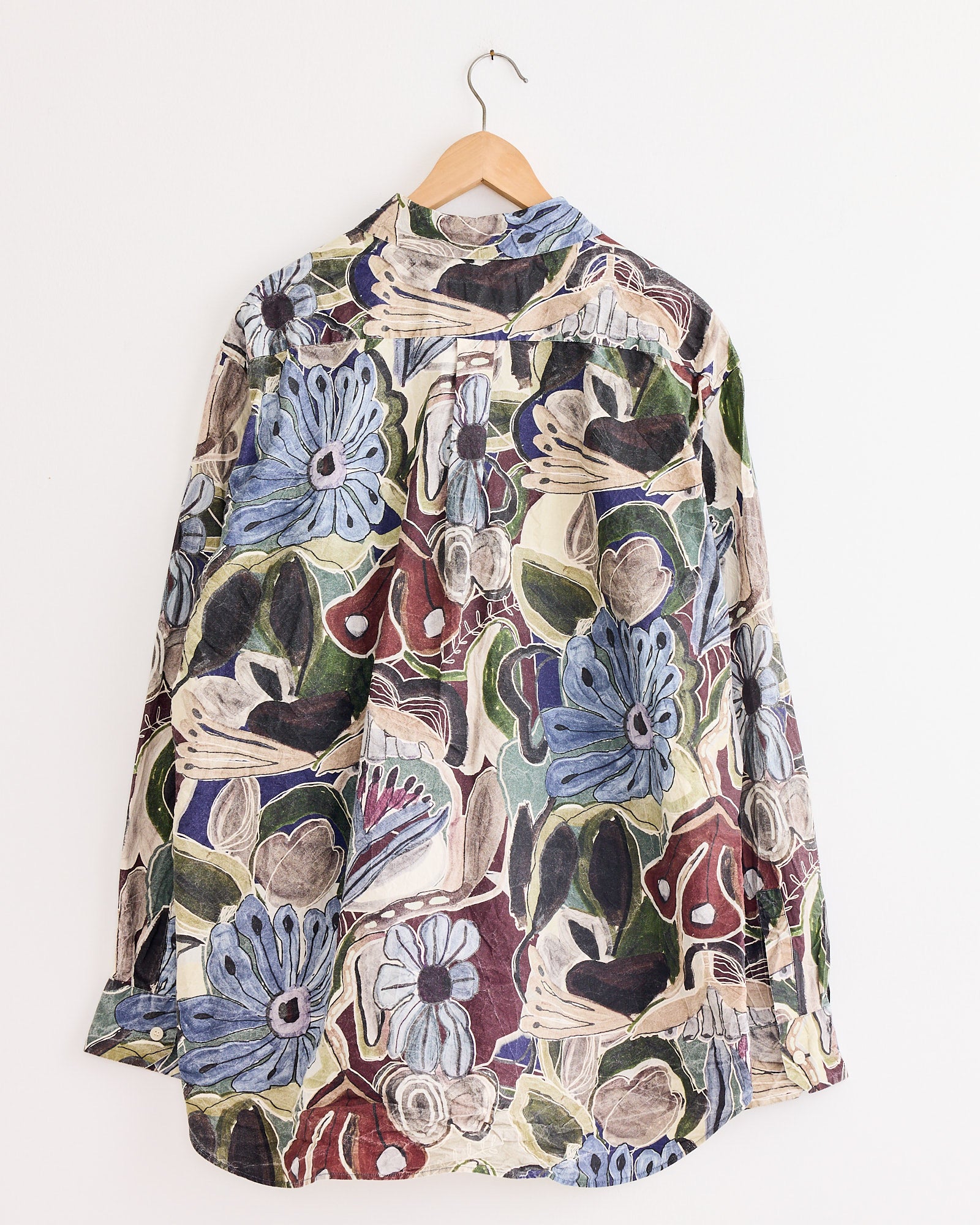 The Casey Casey Big Raccourcie Shirt in Brown, featuring long sleeves and a bold floral pattern in blue, green, brown, and cream, is displayed on a wooden hanger against a plain white wall.