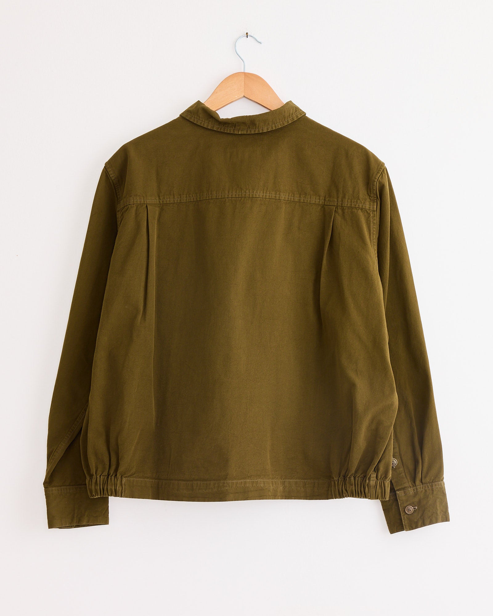 A Casey Casey Fabu Shirt in Green Lake hangs on a wooden hanger against a white background. Made from 100% cotton, it features a collar, long sleeves with buttoned cuffs, and a relaxed fit with subtle gathering at the hem.
