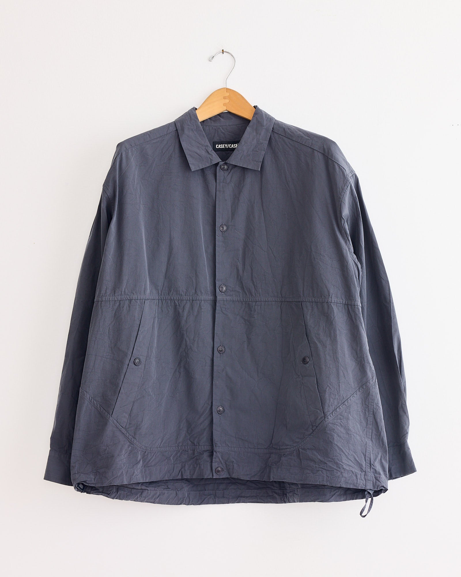 A long-sleeve, blue "Mika Shirt in Blu Skull" by Casey Casey, featuring a collar, front pockets, and a slightly wrinkled texture, hangs on a wooden hanger against a white wall.