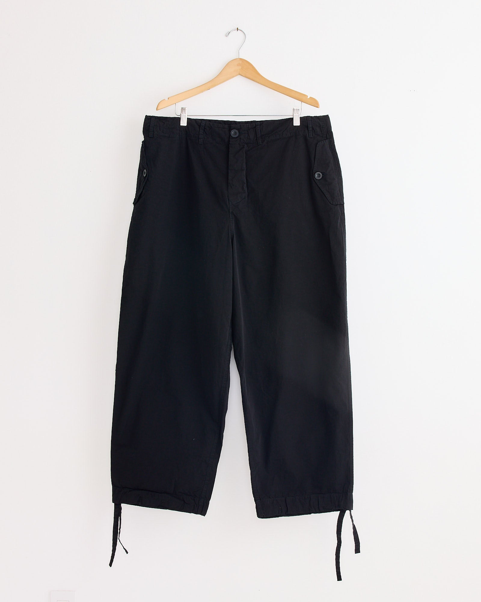 The Casey Casey Abel Pant in Pitch Black hangs on a wooden hanger against a white wall. This relaxed, loose-fit style features side pockets with buttoned flaps and adjustable drawstring cuffs for an effortless look.