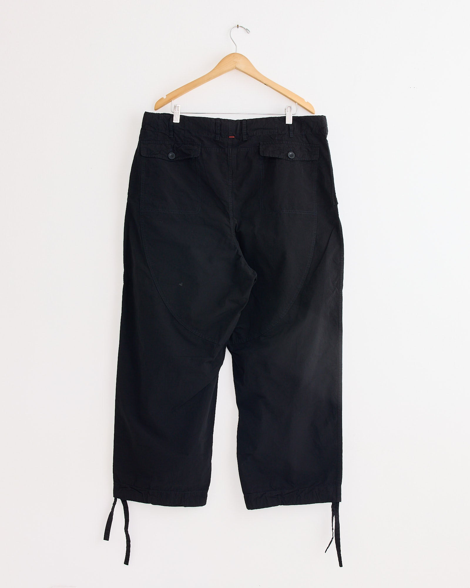 A pair of Casey Casey Abel Pant in Pitch Black hangs on a wooden hanger against a white wall, shown from the back. These pants feature buttoned back pockets and relaxed drawstring cuffs.