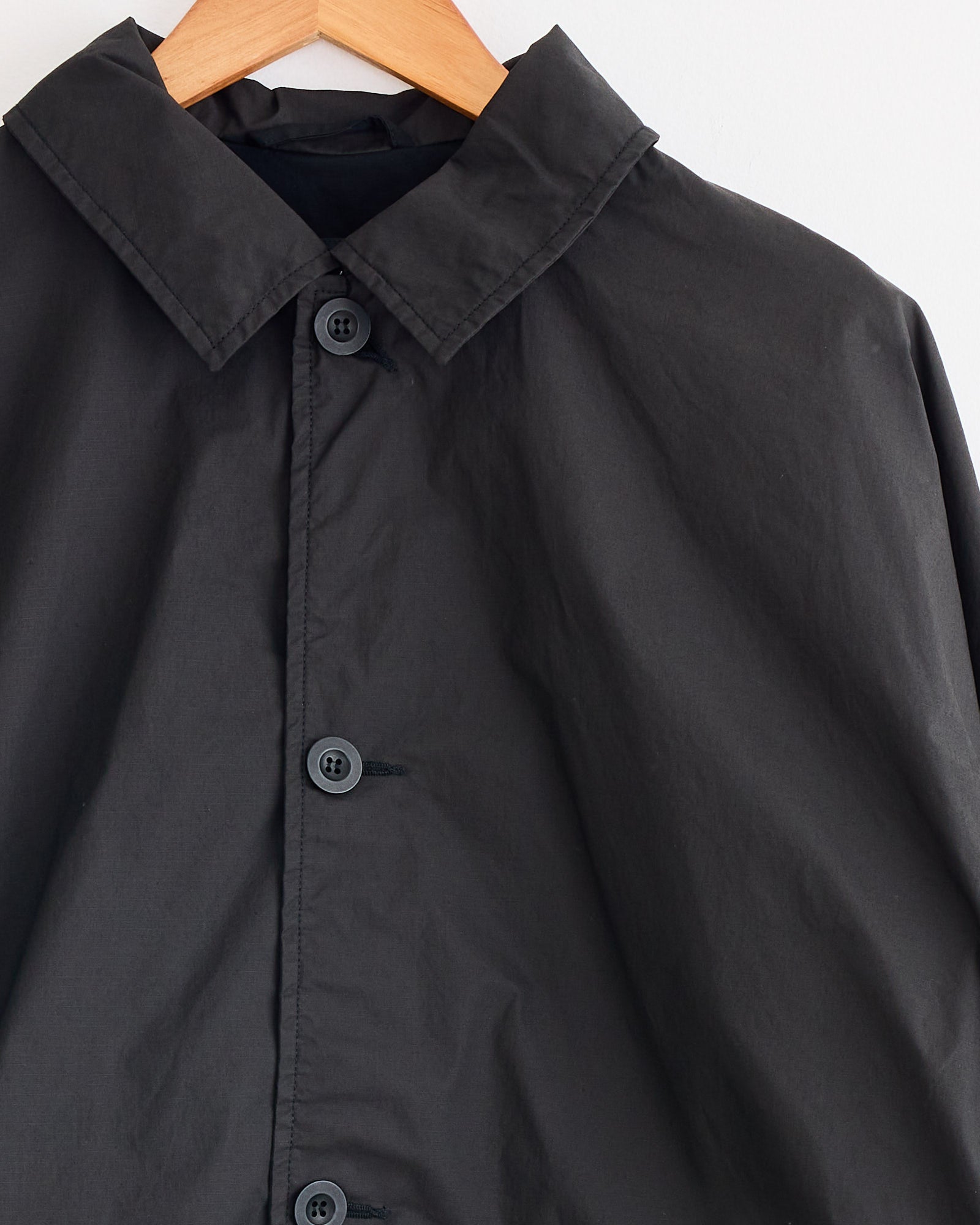 A close-up of the Casey Casey Peche Blouson in Rye, a button-up coat with a collar, hanging on a wooden hanger against a light-colored background.