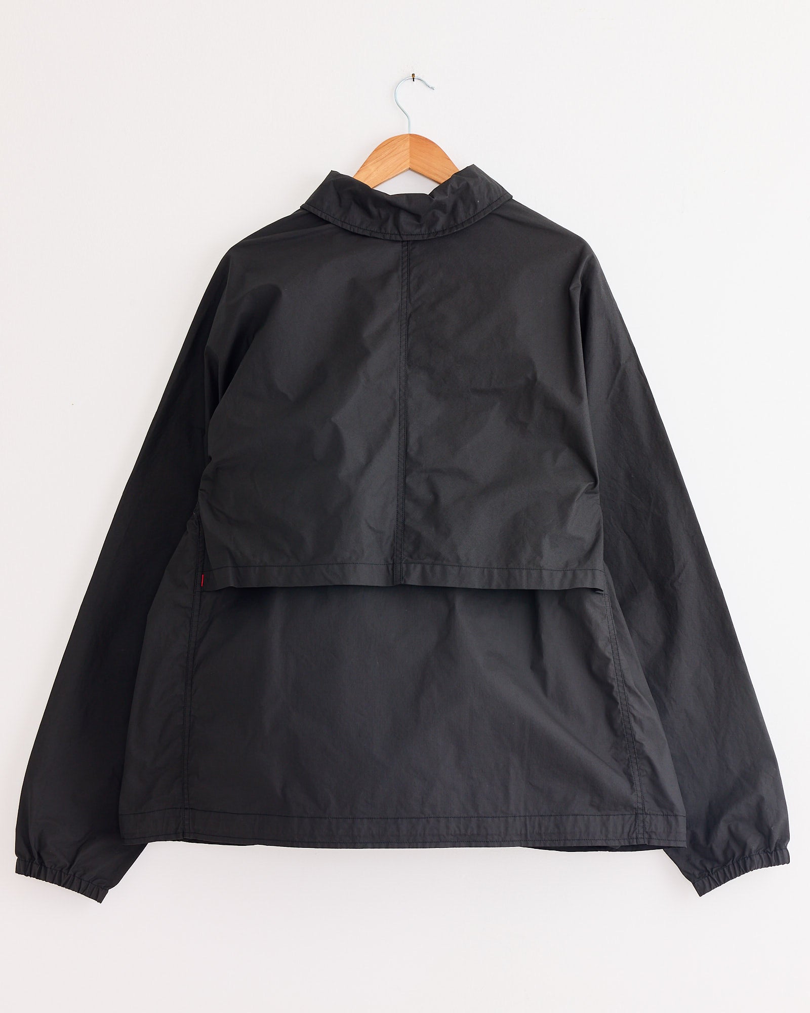 A Casey Casey Peche Blouson in Rye, a waxed cotton jacket with a high collar and chest flap, hangs on a wooden hanger against a plain white wall.