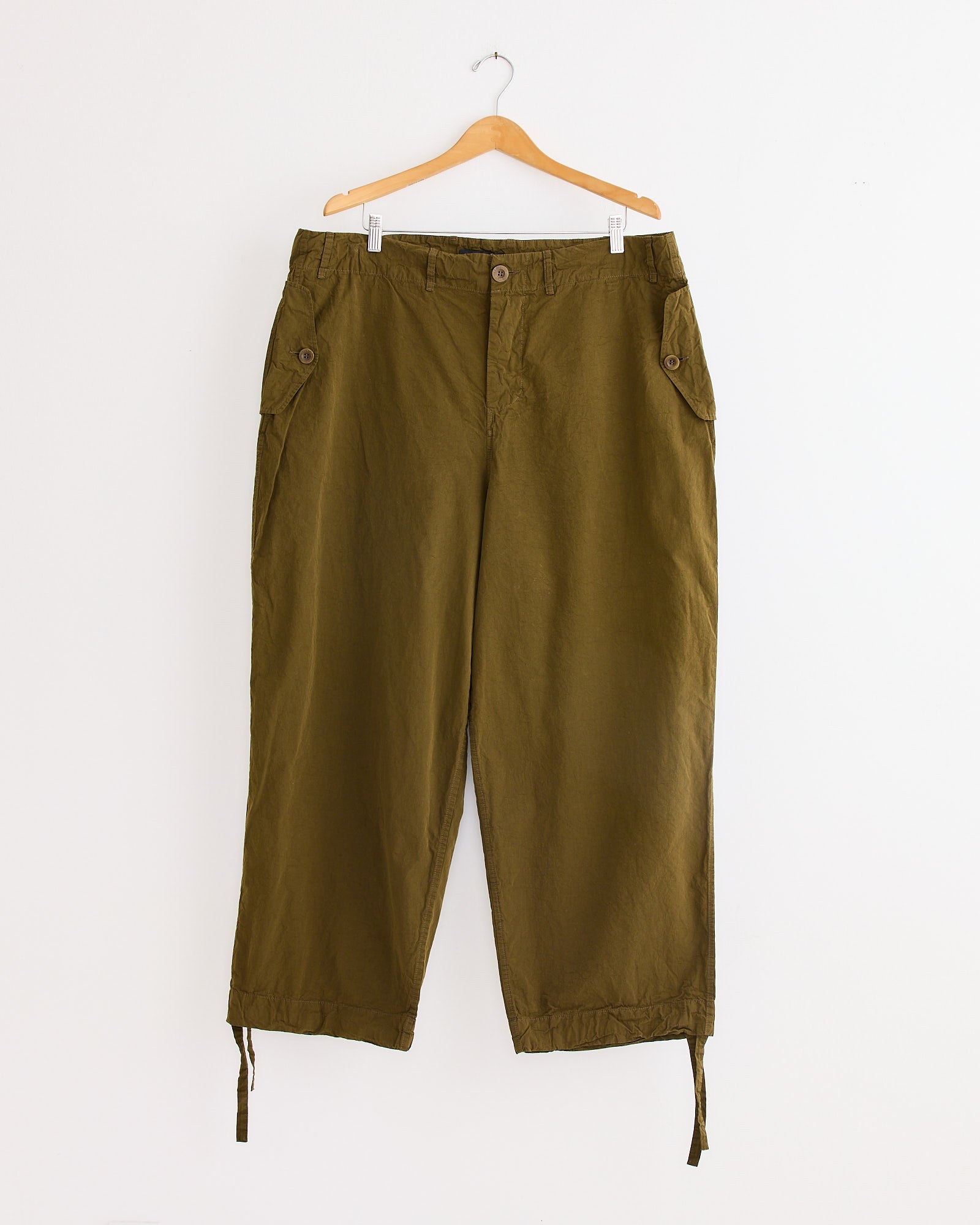 The Casey Casey Abel Pant in Green Lake is a relaxed paper cotton drawstring pant with button and zip closure, side flap pockets with buttons, and adjustable hems, shown on a wooden hanger against a white background.