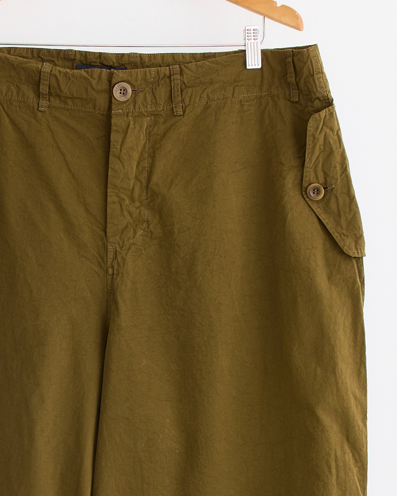 Close-up of the Casey Casey Abel Pant in Green Lake, made from Paper Cotton, hanging on a wooden hanger and showing the waistband, button closure, belt loops, and a side flap pocket with button.