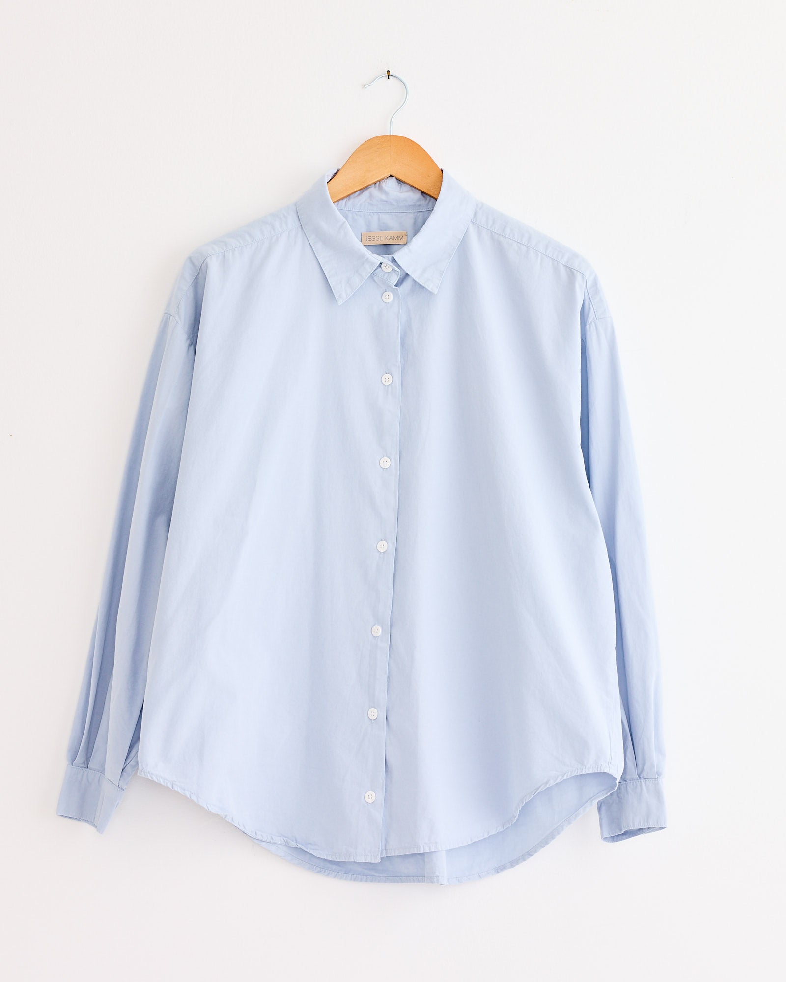 The Jesse Kamm Alberta Shirt in Smokey Blue, a relaxed fit long-sleeve organic cotton button-up, hangs on a wooden hanger against a plain white wall.