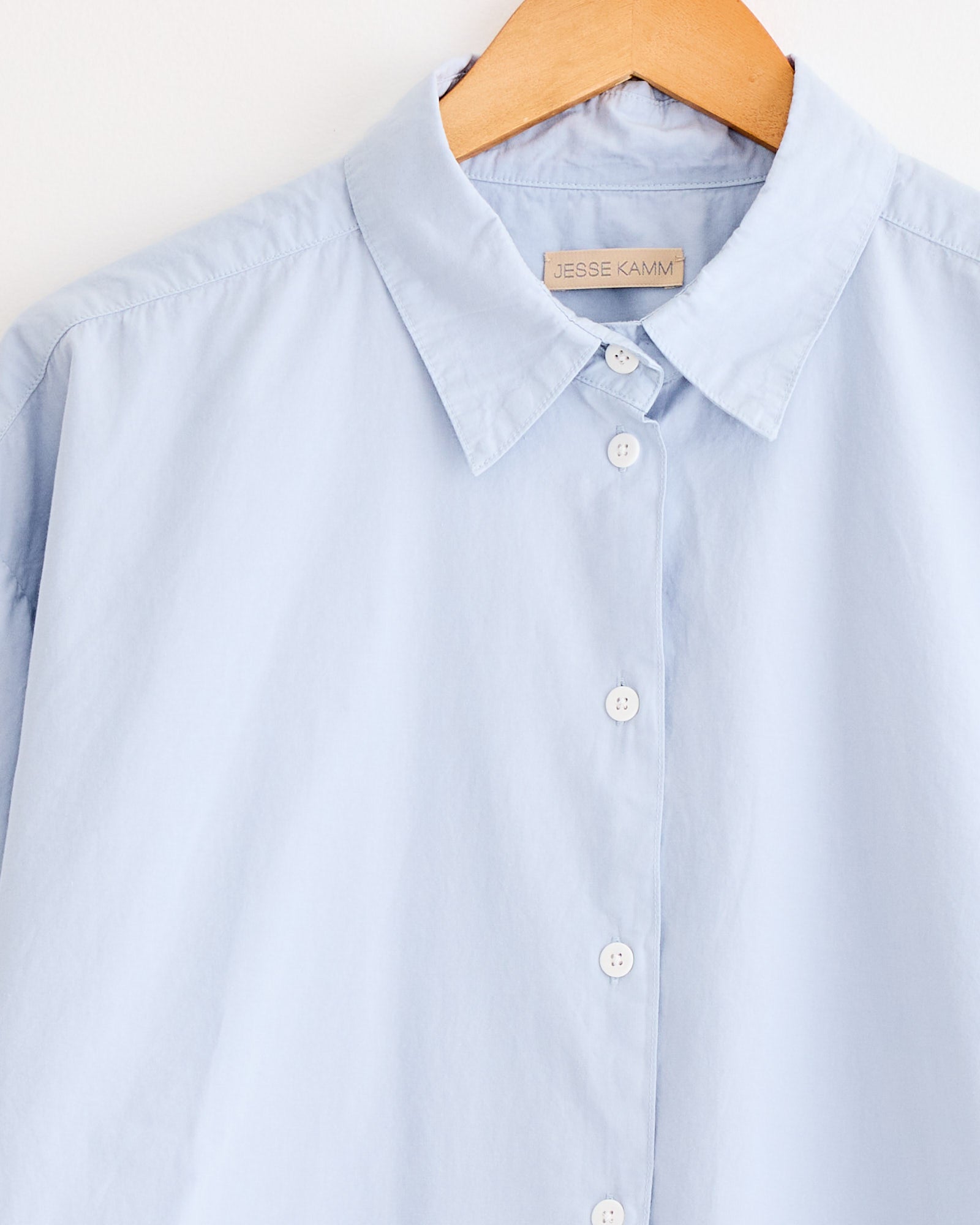 The Jesse Kamm Alberta Shirt in Smokey Blue is a relaxed-fit button-up with a classic collar and white buttons, shown on a wooden hanger with the JESSE KAMM label visible inside the collar.