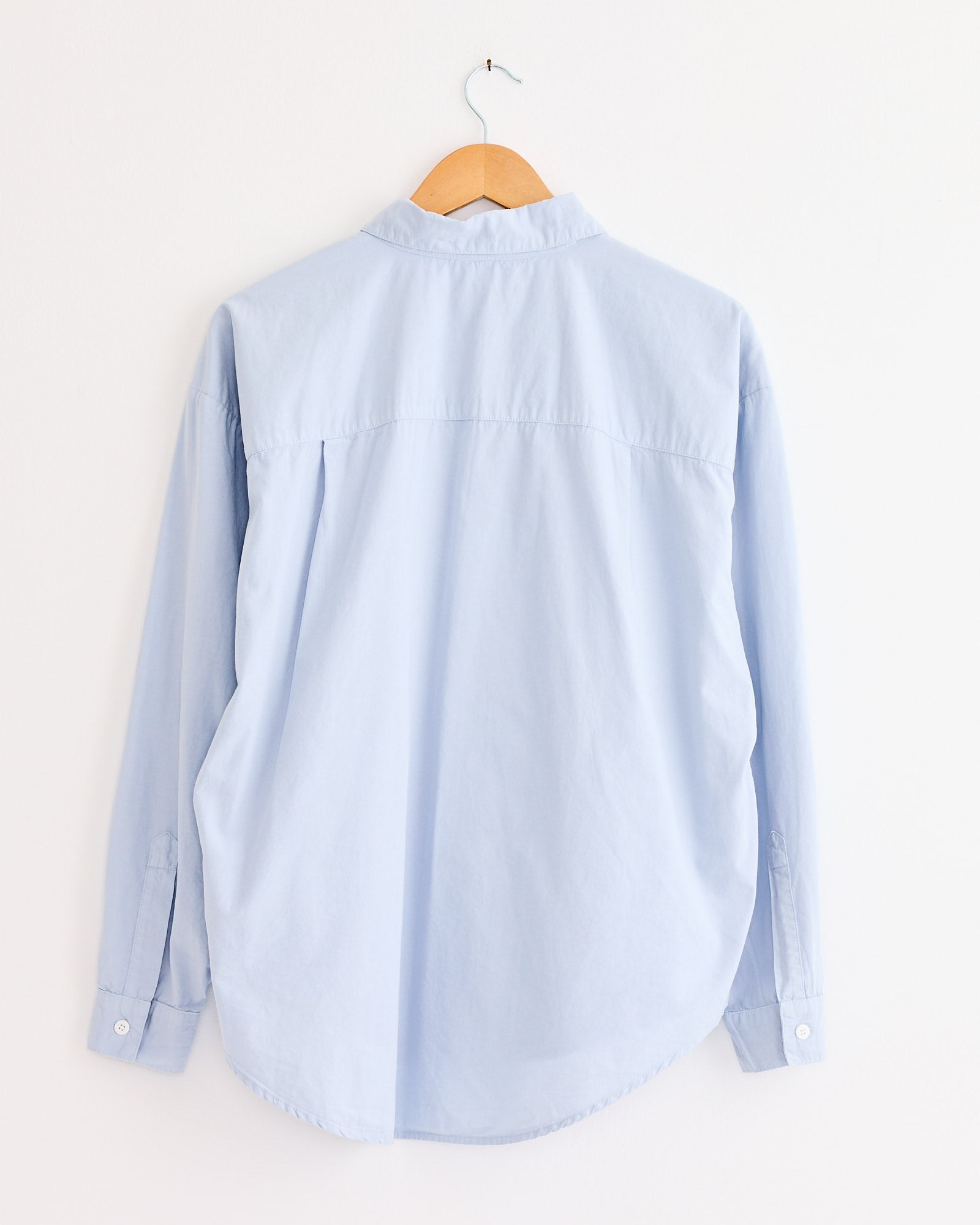 The Jesse Kamm Alberta Shirt in Smokey Blue, made from organic cotton with long sleeves, is shown from the back on a wooden hanger against a plain white wall.