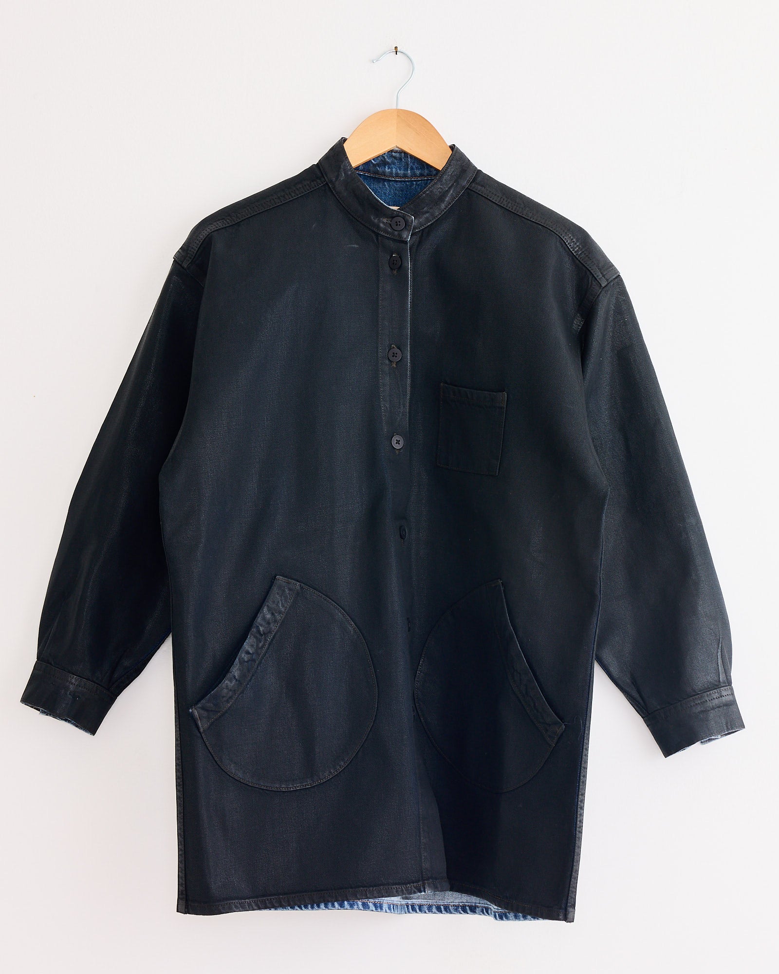 The Jesse Kamm Band Collar Okuda Jacket in Black Lacquer, made from premium Japanese denim, features a mandarin collar, chest pocket, and two large rounded front pockets. Shown hanging on a wooden hanger against a white wall.