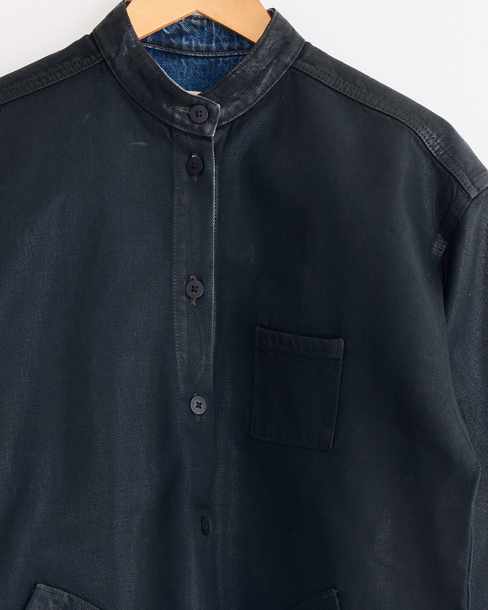 The Jesse Kamm Band Collar Okuda Jacket in Black Lacquer, crafted from Japanese denim with a collarless button-up design and chest pocket, is displayed on a hanger against a plain white background.