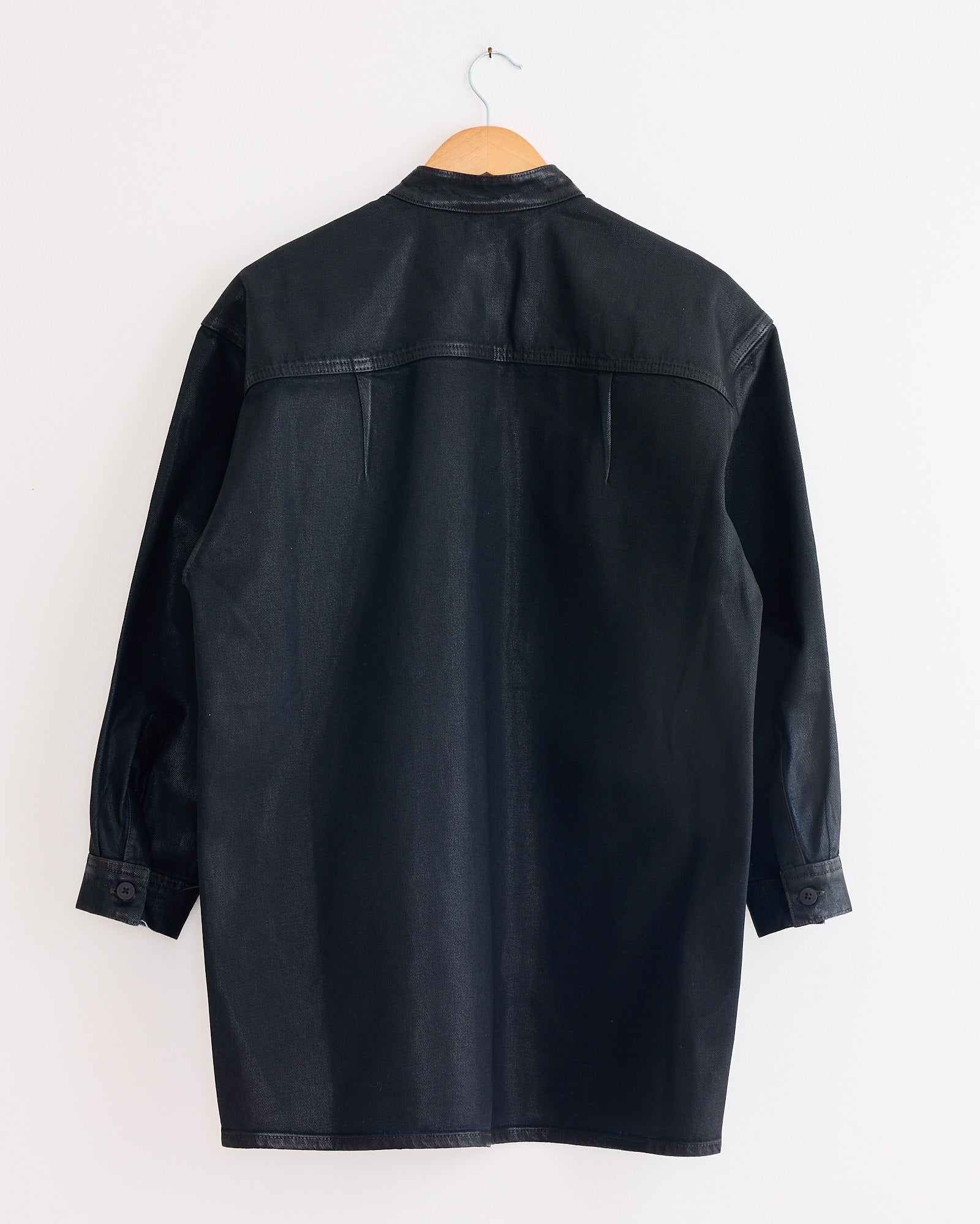 The Jesse Kamm Band Collar Okuda Jacket in Black Lacquer hangs on a wooden hanger against a plain white wall, shown from the back with visible seams and buttoned cuffs.