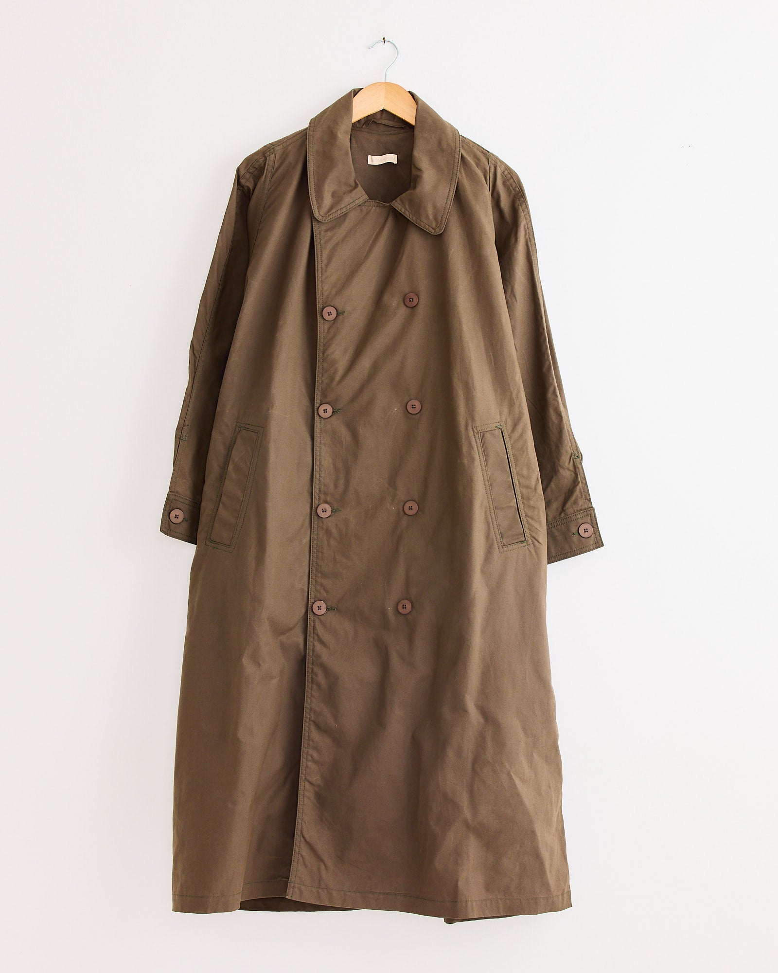 The Jesse Kamm Car Coat in Loam is a brown deadstock coat with wide lapels, front buttons, and two large pockets, displayed on a wooden hanger against a plain white wall.
