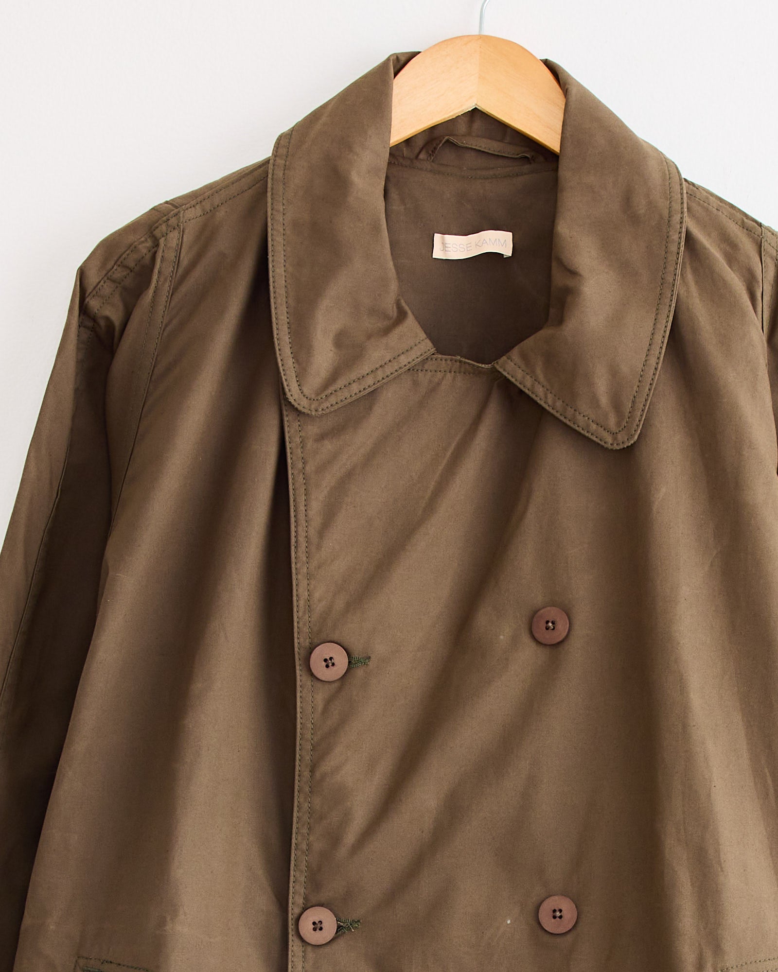 The Car Coat in Loam by Jesse Kamm, a brown double-breasted coat with wide lapels and four visible buttons, hangs on a wooden hanger against a white wall, featuring a beige tag sewn inside the collar.