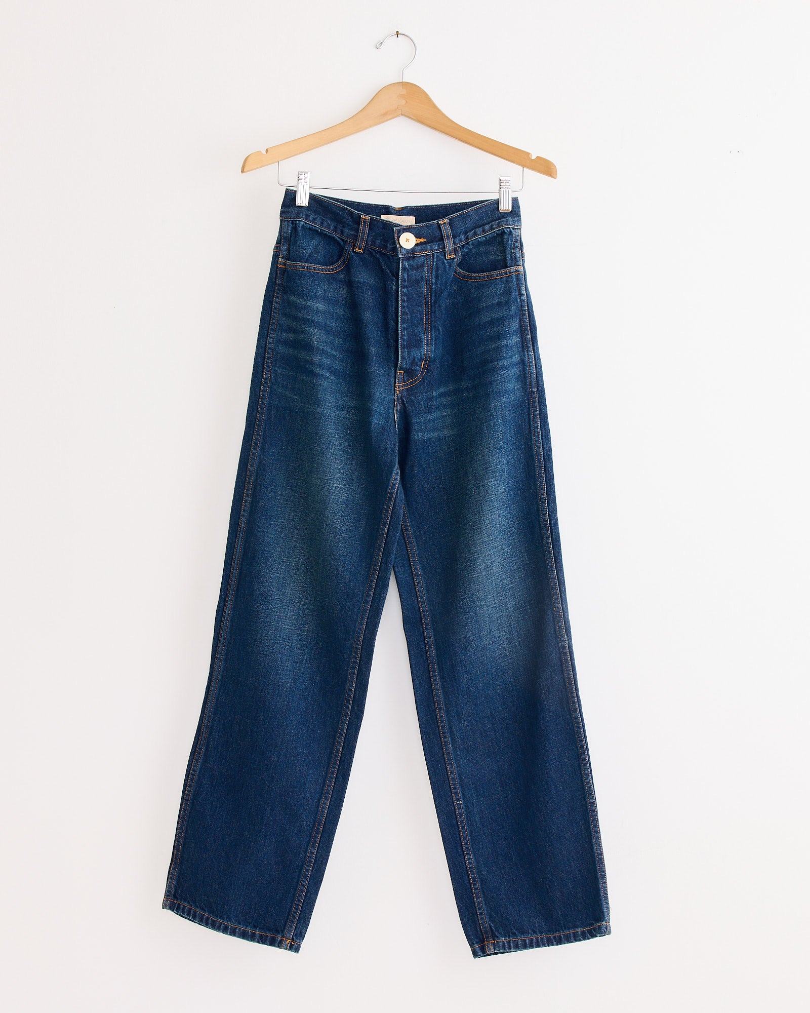 The 225 Pant in Medium Roast by Jesse Kamm, a high-waisted, wide-leg blue jean crafted from regenerative Japanese denim, hangs on a wooden hanger against a plain white wall.