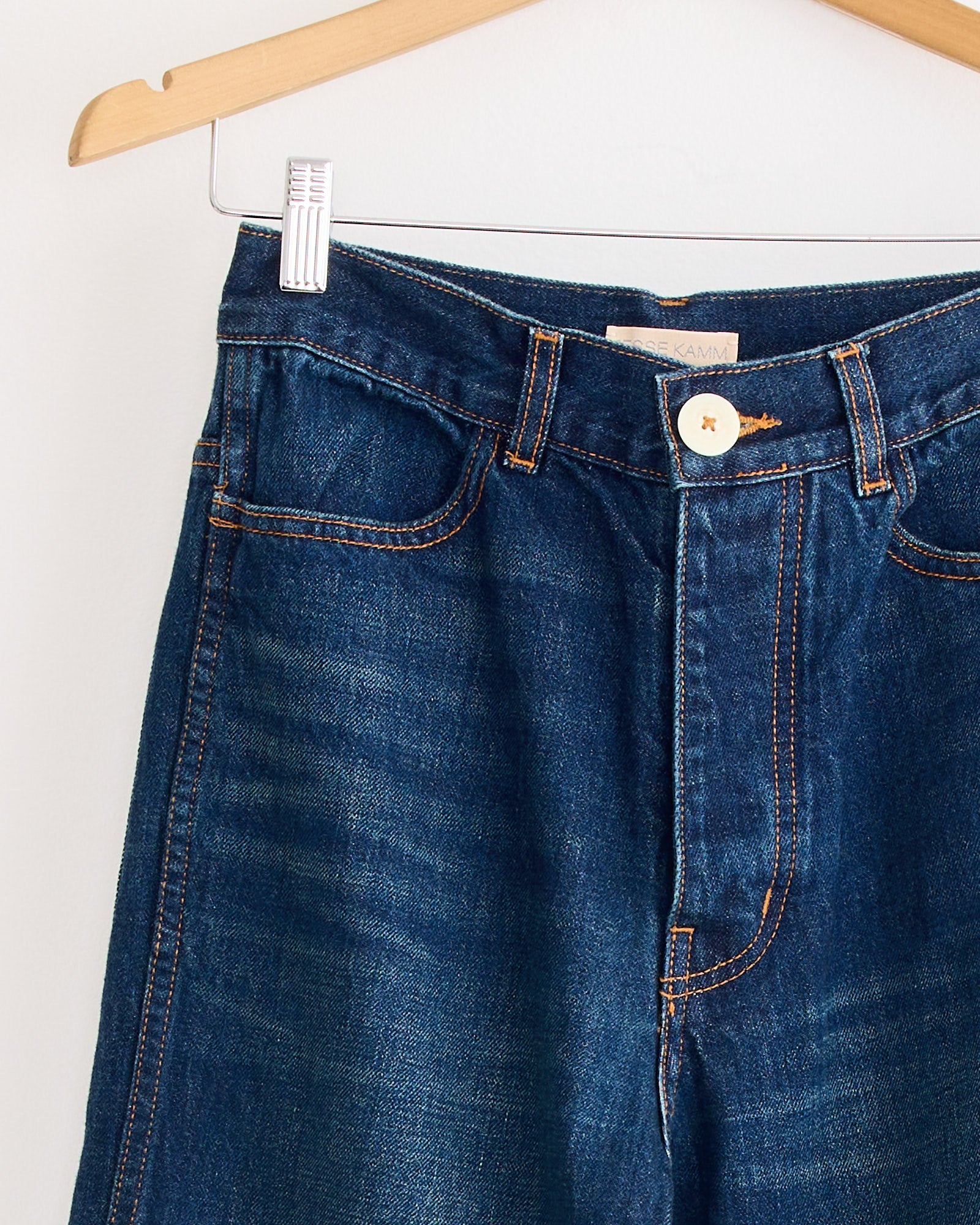 A close-up of The 225 Pant in Medium Roast by Jesse Kamm, featuring a high waist, waistband, button, front pockets, and orange stitching, crafted from regenerative Japanese denim and hanging on a wooden hanger.