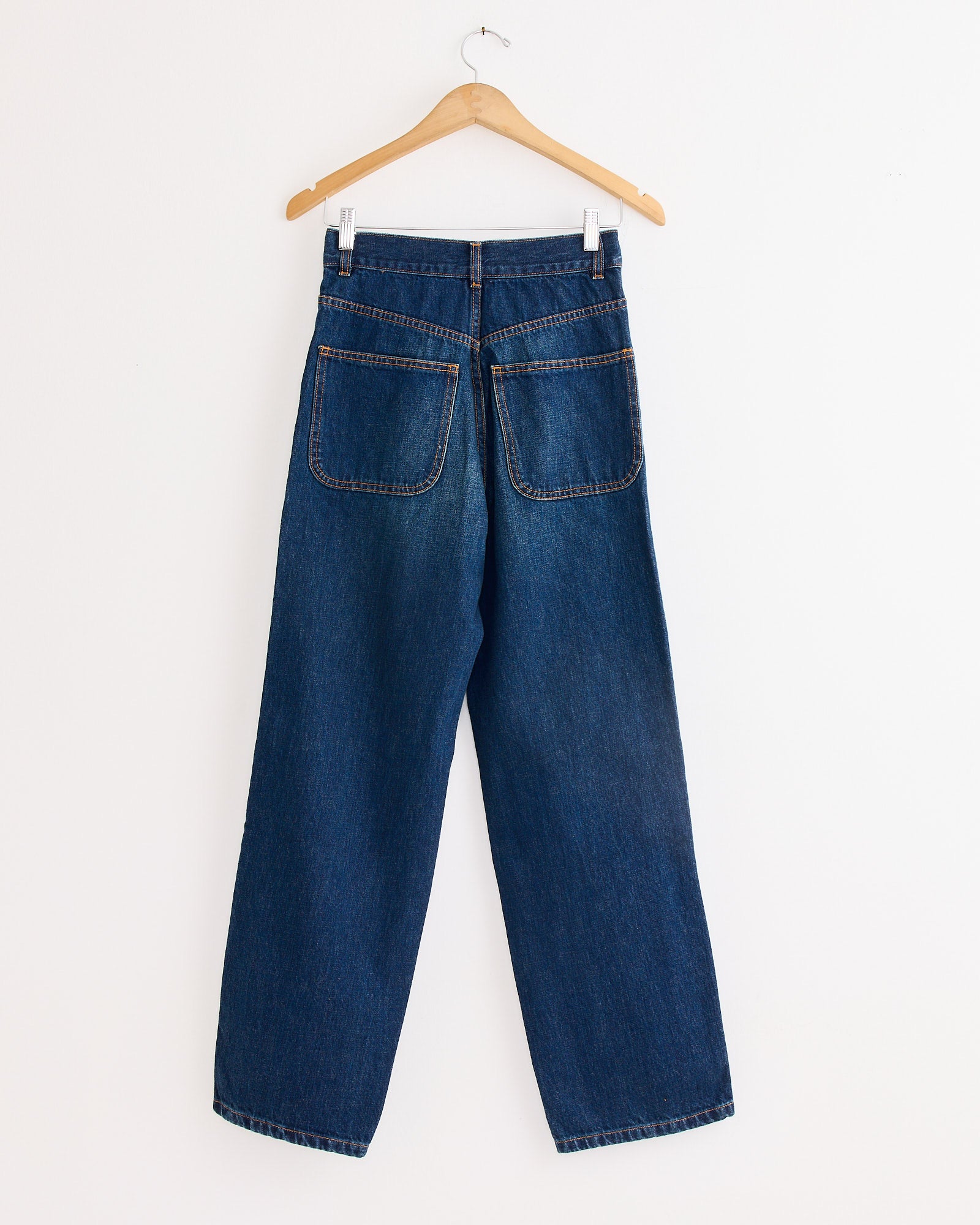 The 225 Pant in Medium Roast by Jesse Kamm hangs on a wooden hanger against a white wall, shown from the back with two large rear pockets and wide, straight legs in dark blue denim.