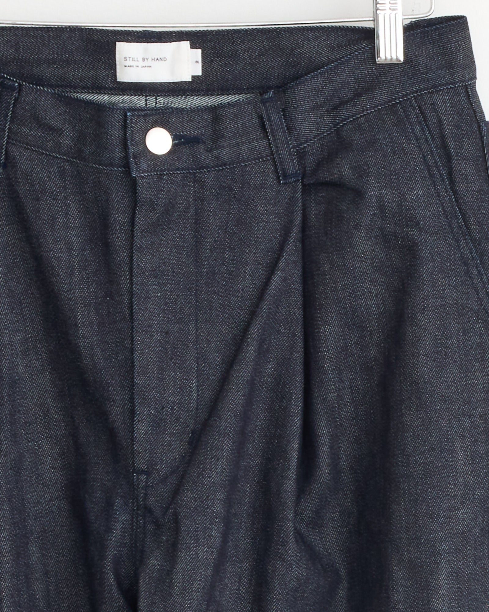 The 225 Pant in Medium Roast by Jesse Kamm features a pleated front, silver button closure, and belt loops. A white label is sewn inside the waistband. The trousers are displayed on a metallic clip hanger.