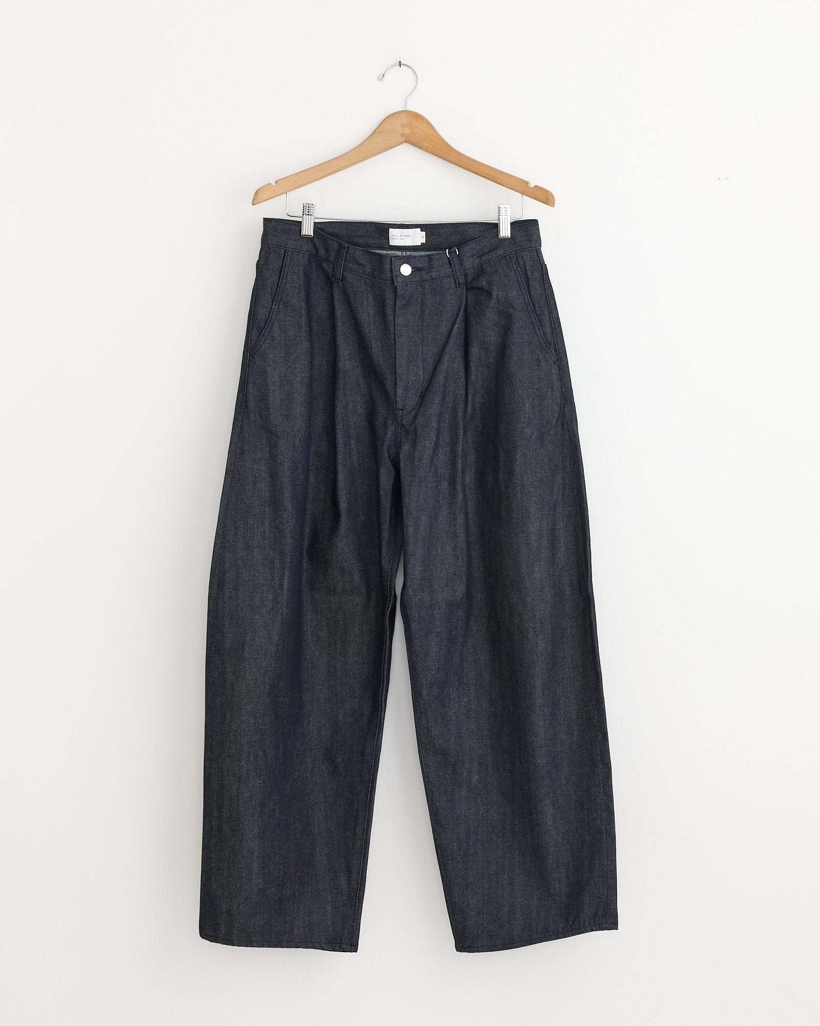 The 225 Pant in Medium Roast by Jesse Kamm hangs on a wooden hanger against a plain white background. These wide-leg, high-waist jeans in dark Japanese denim feature front pleats, side pockets, and a button closure.