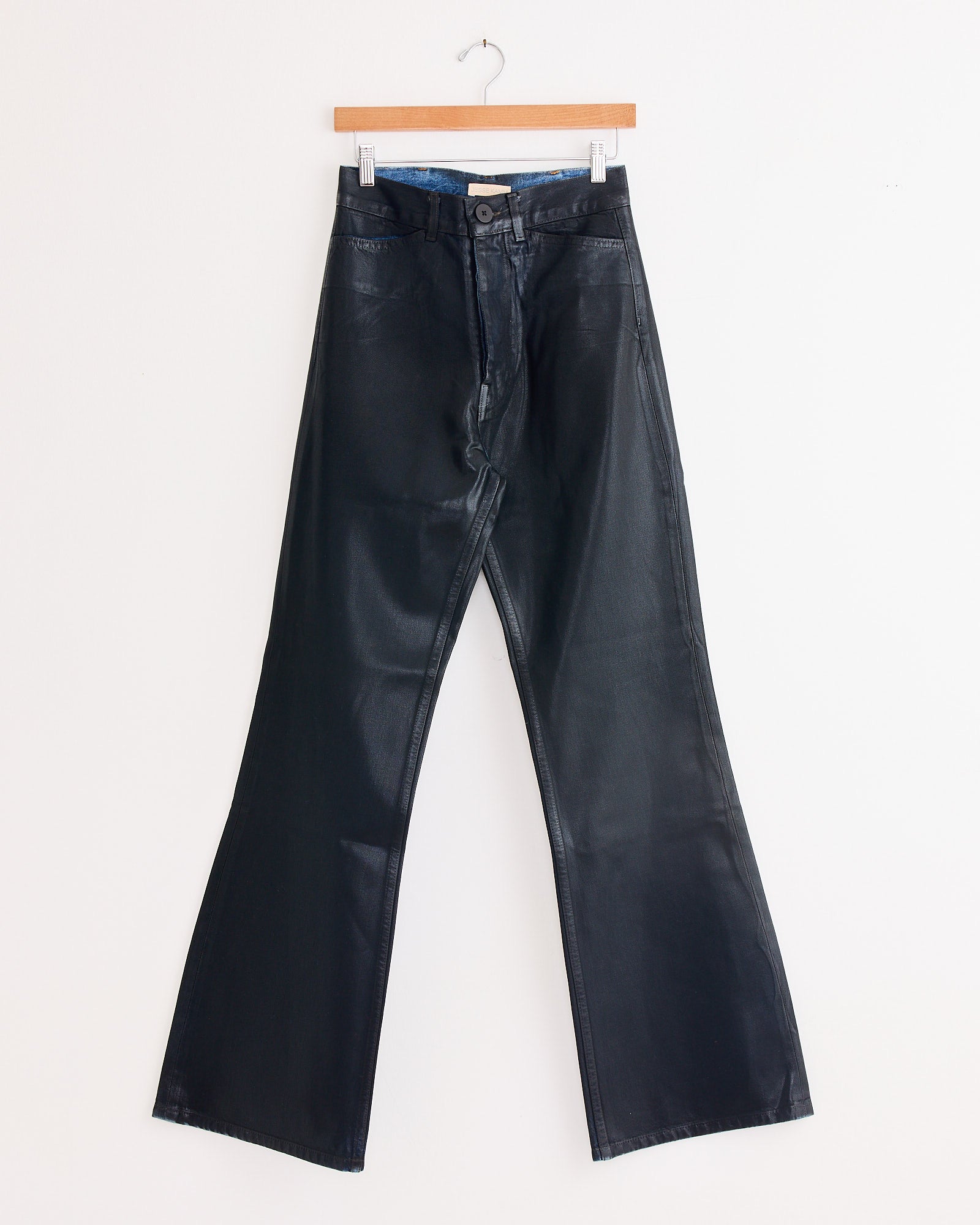 The Les Deux Pant in Black Lacquer by Jesse Kamm hangs on a wooden hanger against a white wall. These high-waisted flared pants feature a front button and five-pocket design, evoking the brand’s classic style.