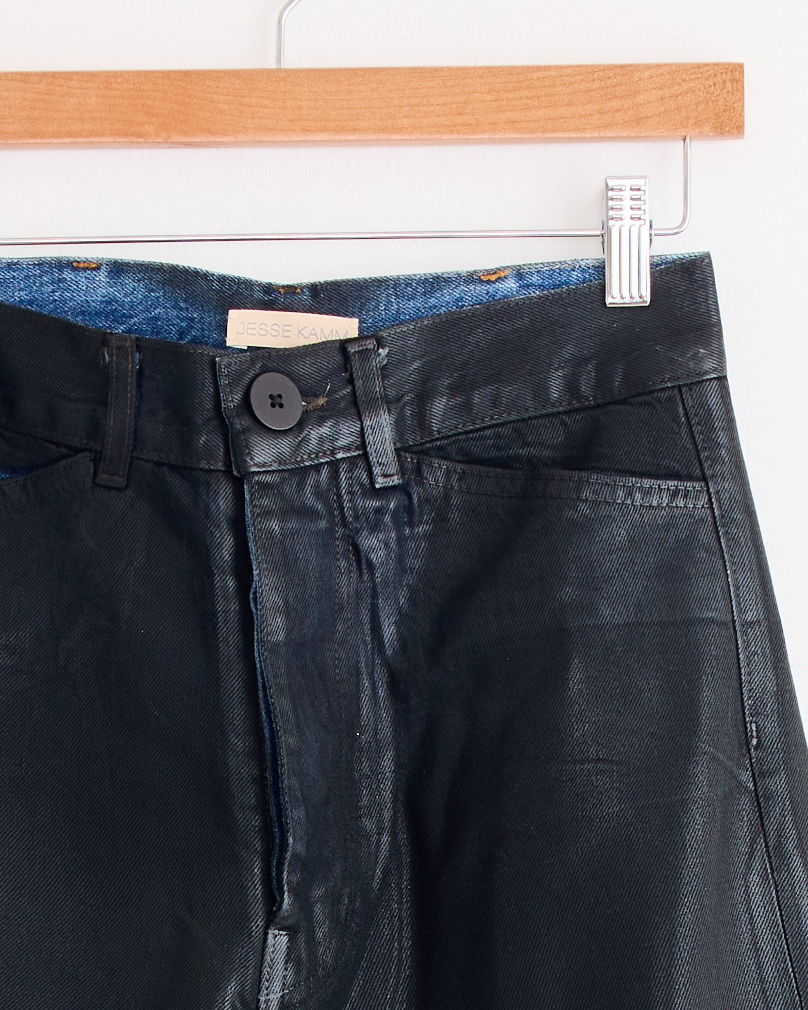 Close-up of the Jesse Kamm Les Deux Pant in Black Lacquer hanging on a wooden hanger, with blue denim lining and a visible brand label inside the high waisted, flared silhouette.
