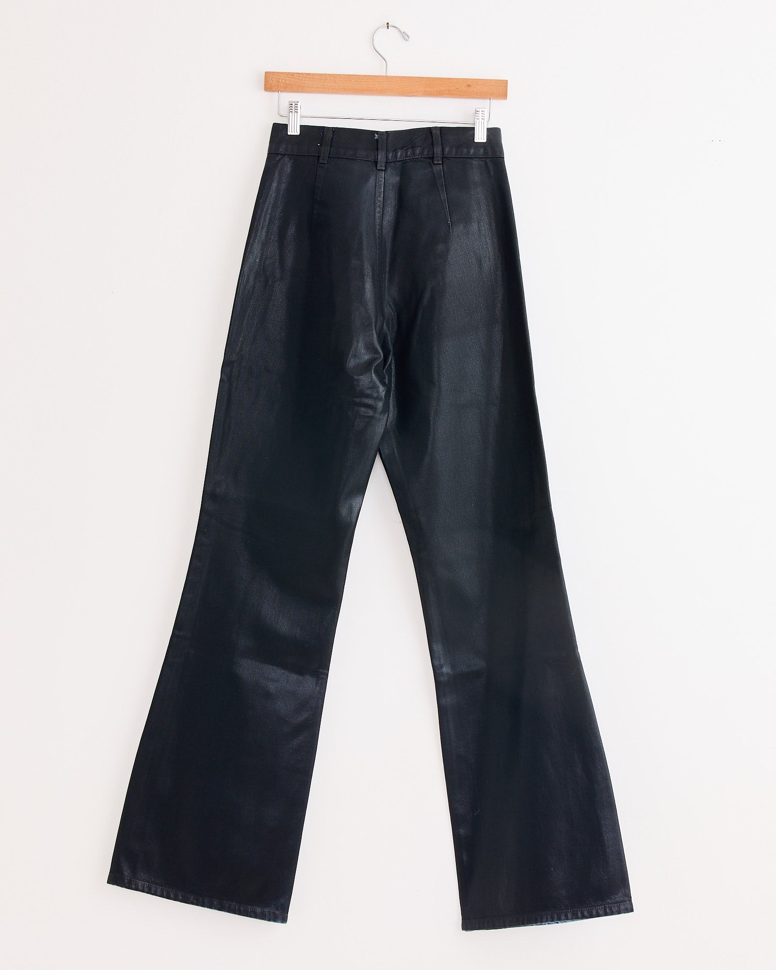 The Les Deux Pant in Black Lacquer by Jesse Kamm—high-waisted, wide-leg faux leather—hangs on a wooden hanger against a plain white background.