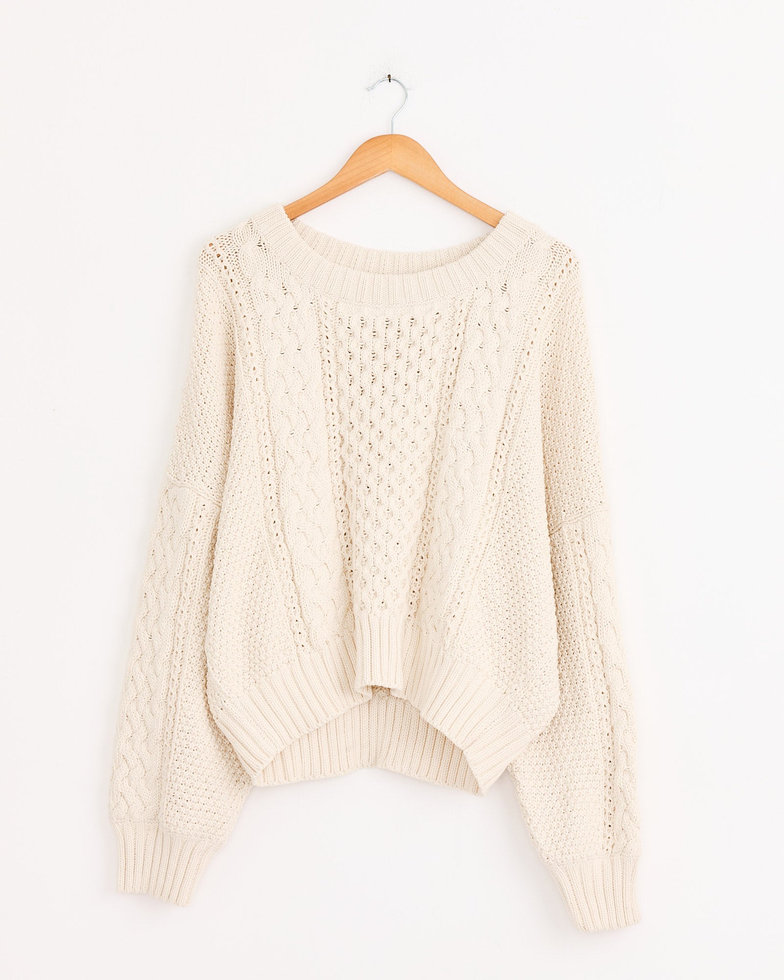 The Shaina Mote Fisherman Sweater in Natural, a cream cable knit with chunky classic styling, hangs on a wooden hanger against a plain white wall.