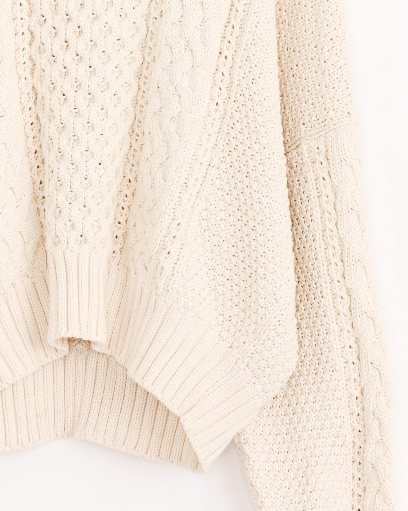 Close-up of the Fisherman Sweater in Natural by Shaina Mote, featuring cream-colored cable knit with varied textured patterns like cables and ribbing, shown hanging against a light background.