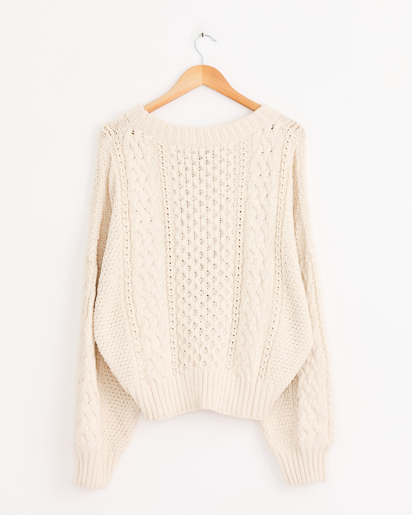 The Fisherman Sweater in Natural by Shaina Mote hangs on a wooden hanger against a white wall, showcasing its cream cable knit, textured patterns, long sleeves, and ribbed cuffs and hem.