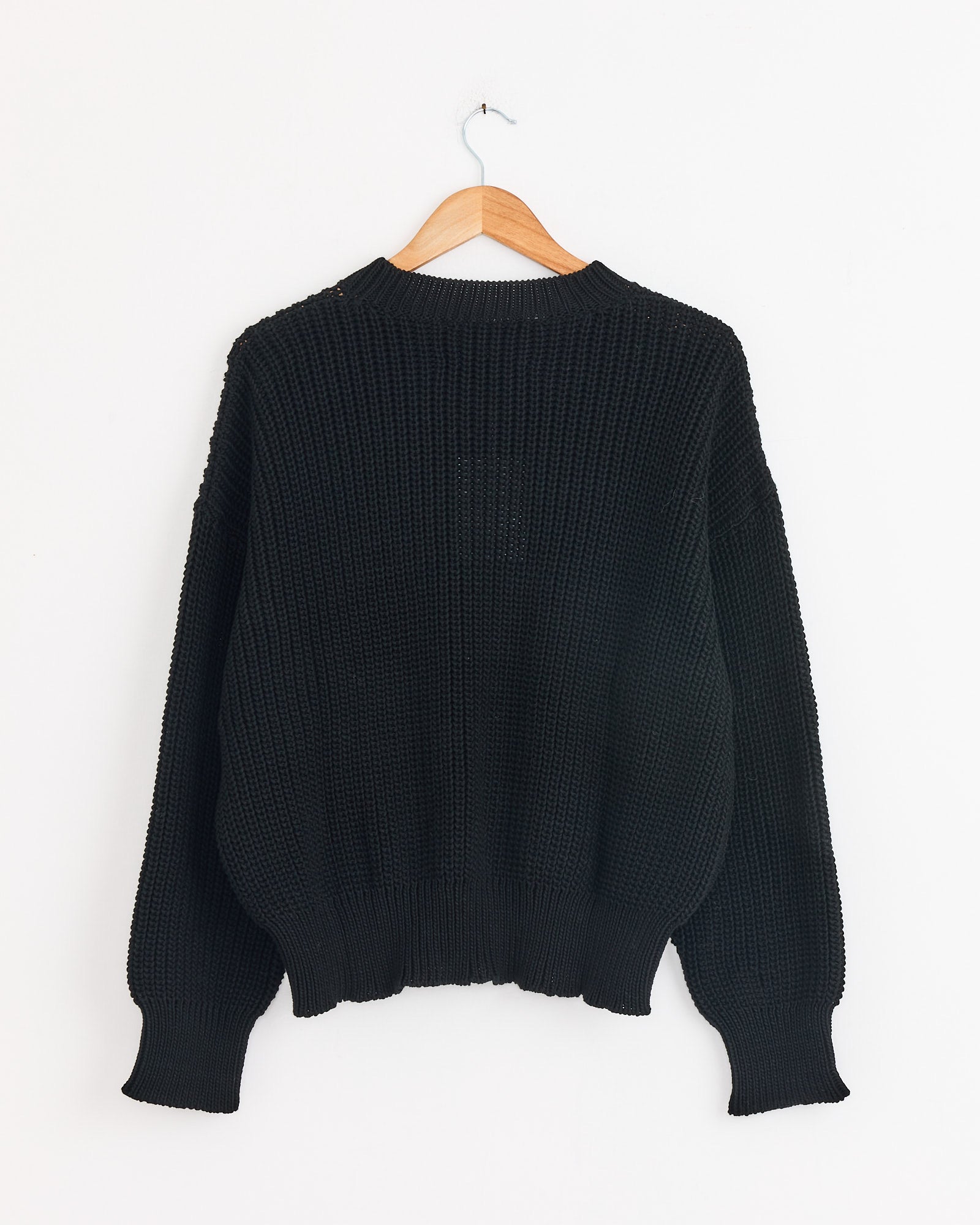 The Shaina Mote Perle Sweater in Onyx, a long-sleeved cropped crewneck, hangs on a wooden hanger against a plain white wall.