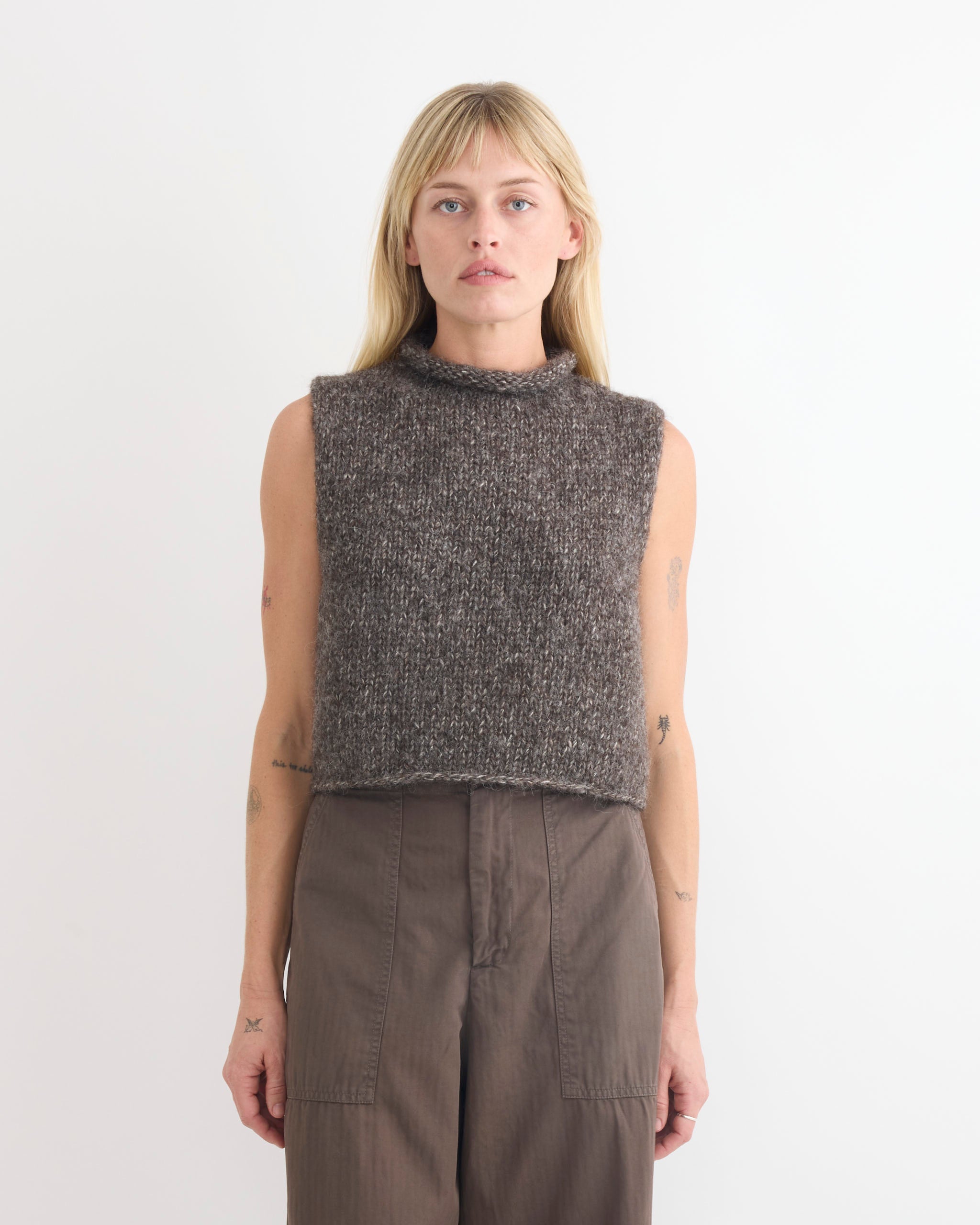 A person with straight blond hair wears the Shaina Mote Roll Vest in Dusk, a sleeveless, high-neck, gray alpaca vest in a cropped fit, paired with high-waisted brown pants against a plain white background.