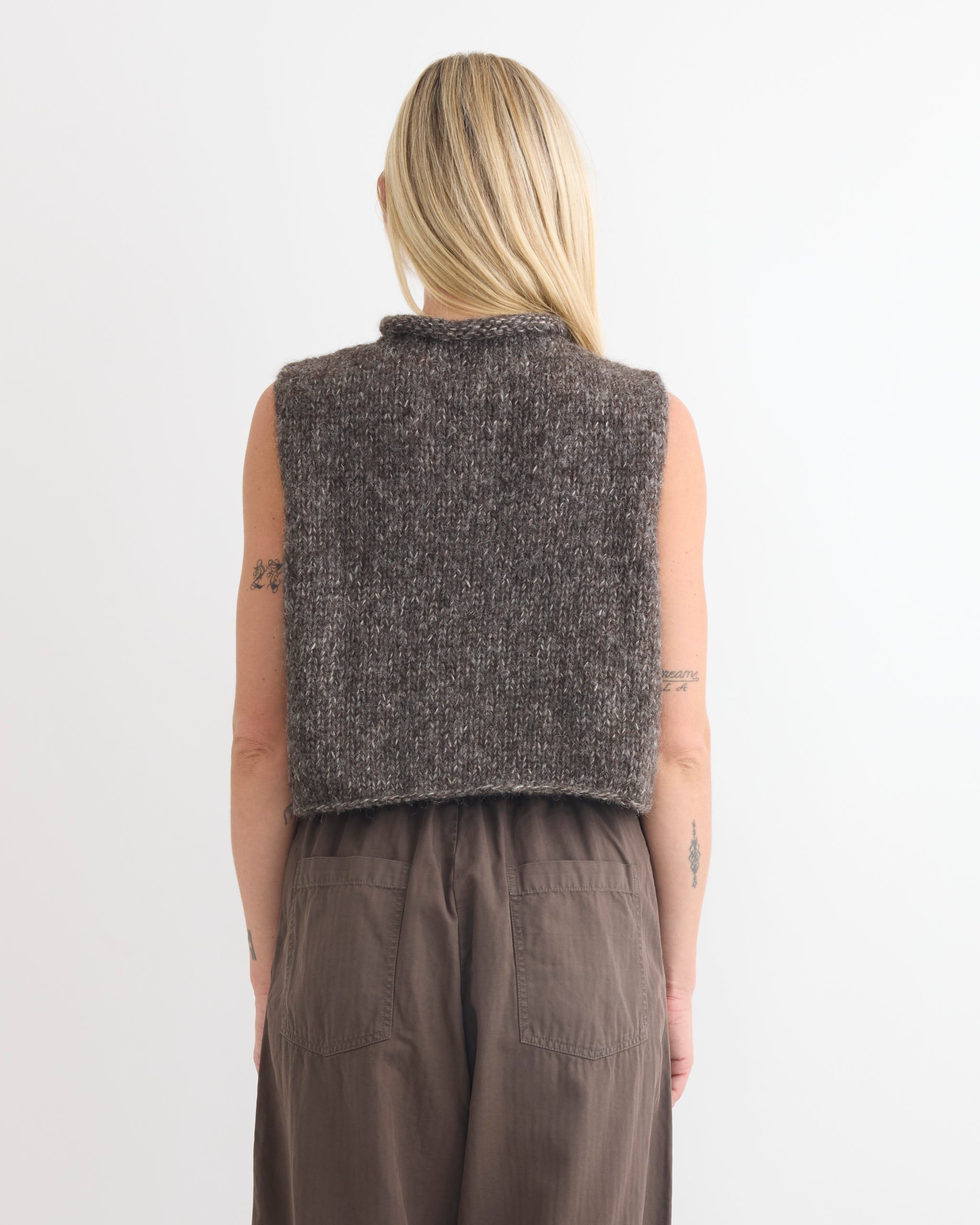 A person with long blonde hair, seen from behind, wears the Shaina Mote Roll Vest in Dusk—a sleeveless, chunky knit alpaca vest with a cropped fit—and high-waisted brown pants against a plain white background.