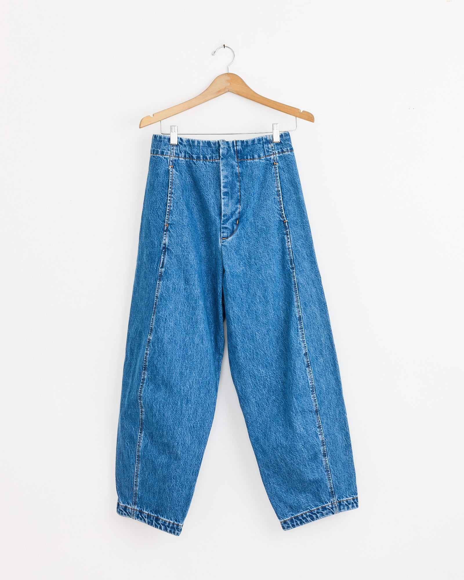 The Shaina Mote Lune Pant in Medium Blue, featuring a high waist and balloon leg silhouette, hangs on a wooden hanger against a plain white wall.