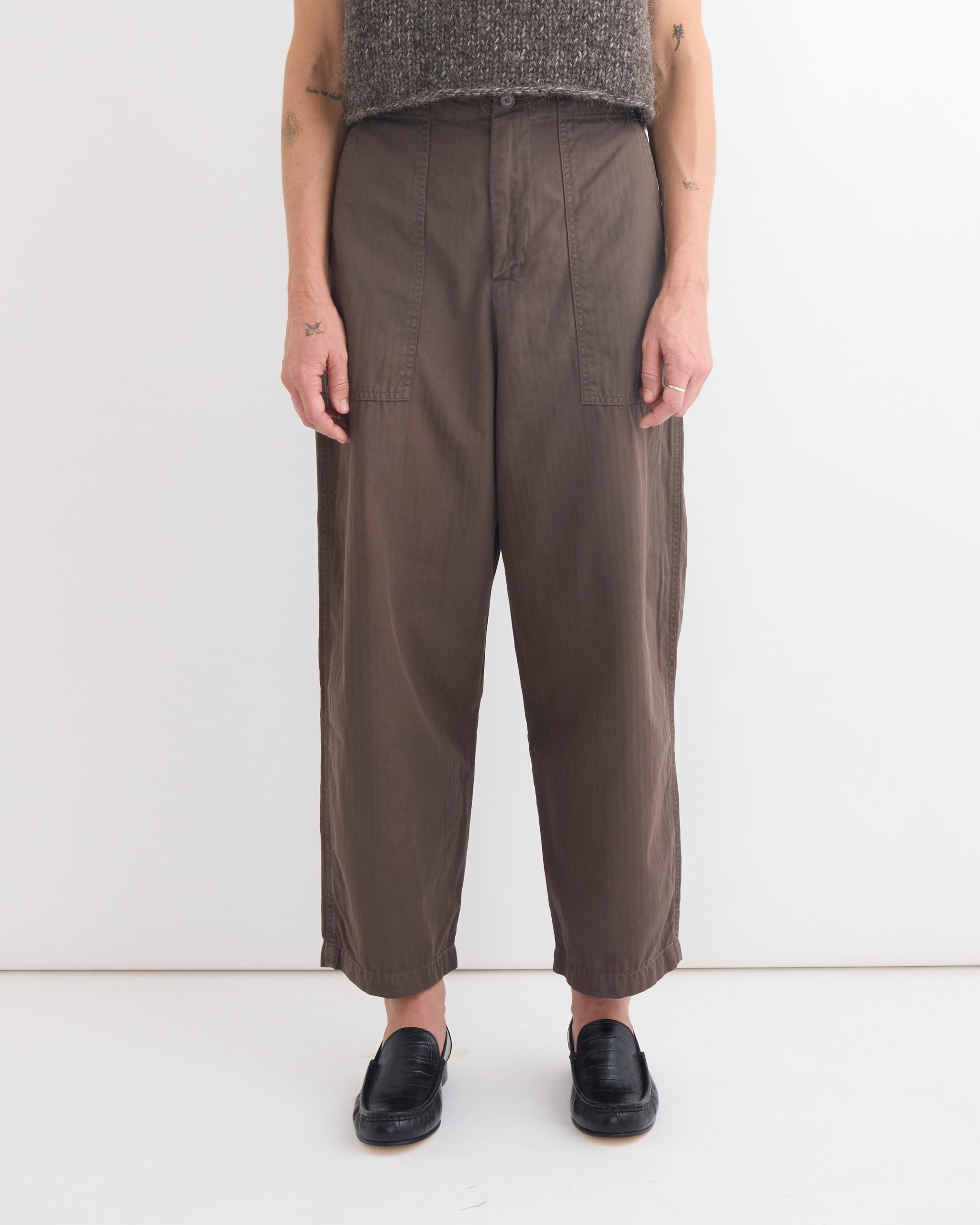 A person stands against a white background wearing Shaina Mote's Painter Pant in Ermine, a textured gray sleeveless top, and black slip-on loafers. Tattoos are visible on both arms for a laid-back, artful look.