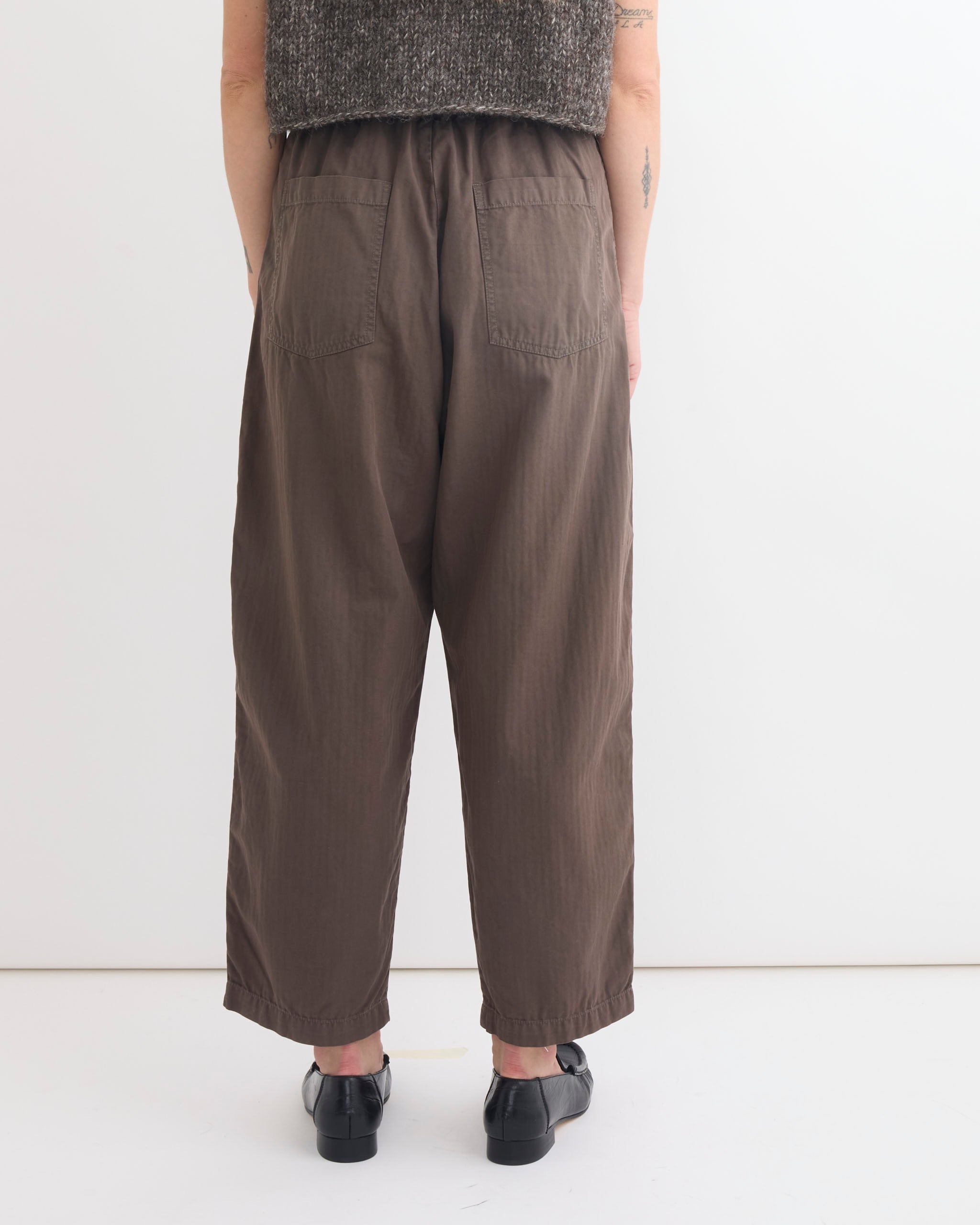 Facing a white wall, only the lower back and legs are visible as the person wears Shaina Mote's Painter Pant in Ermine with back pockets, black shoes, and a sleeveless textured top for a modern, effortless look.