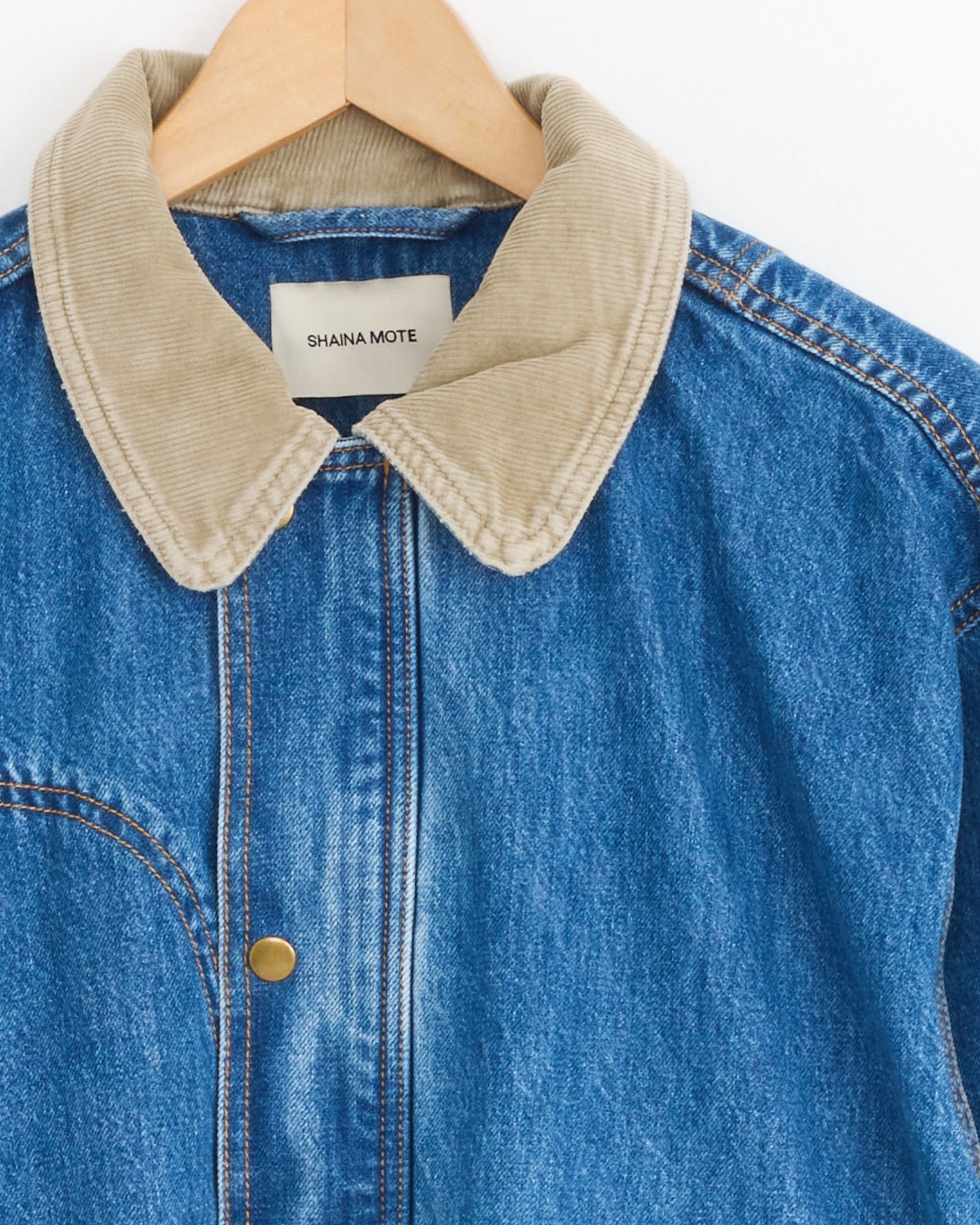 The Town And Country Coat in Medium Blue by Shaina Mote features Italian denim, a tan corduroy collar, brass snap button, contrasting stitching, and is shown on a wooden hanger with the brand label.