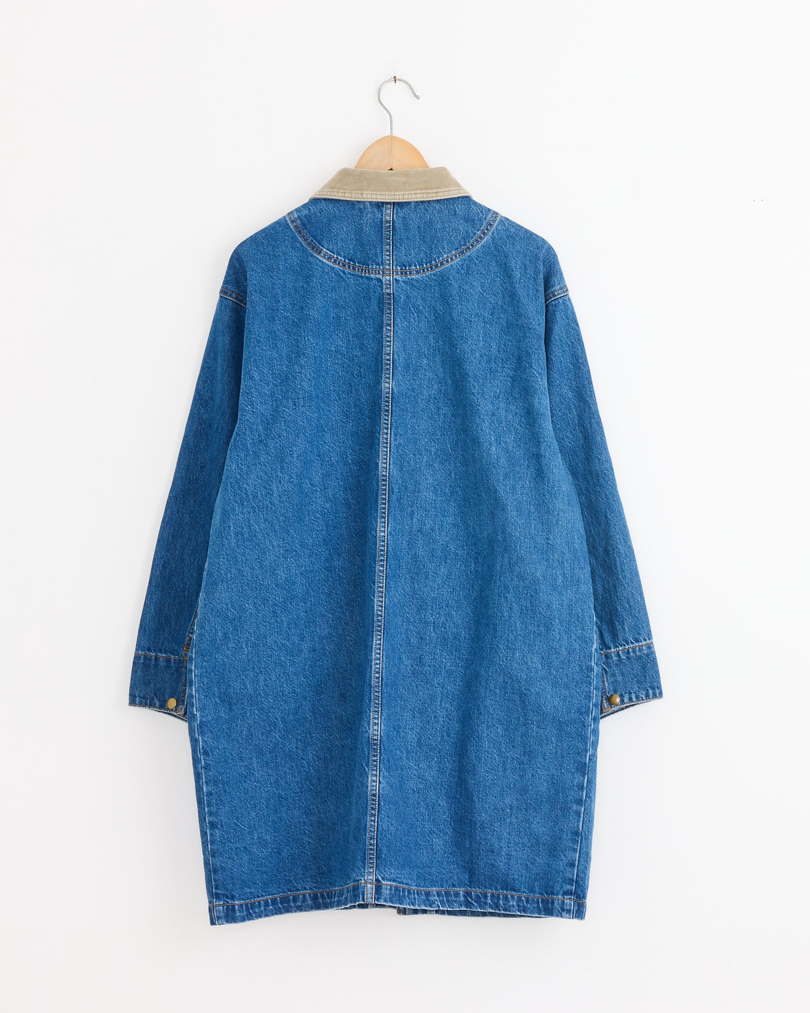 The Shaina Mote Town And Country Coat in Medium Blue, shown from the back on a wooden hanger against a white wall, features long sleeves, Italian denim, a tan collar, and a center seam.