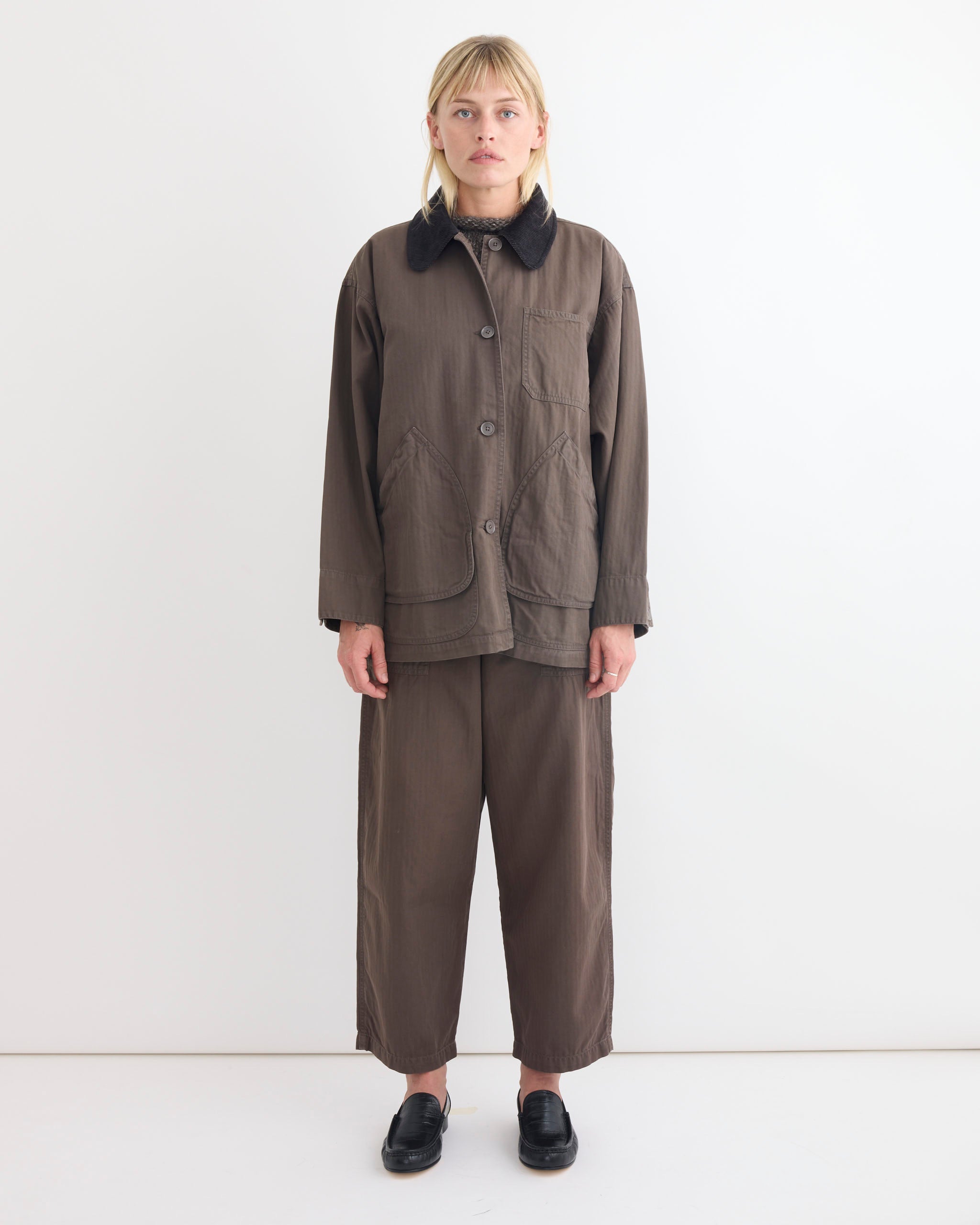 A person stands against a plain white wall, wearing Shaina Mote’s Painter Coat in Ermine—a loose-fitting brown coat with large front pockets and a corduroy collar—paired with matching wide-legged pants and black slip-on shoes.