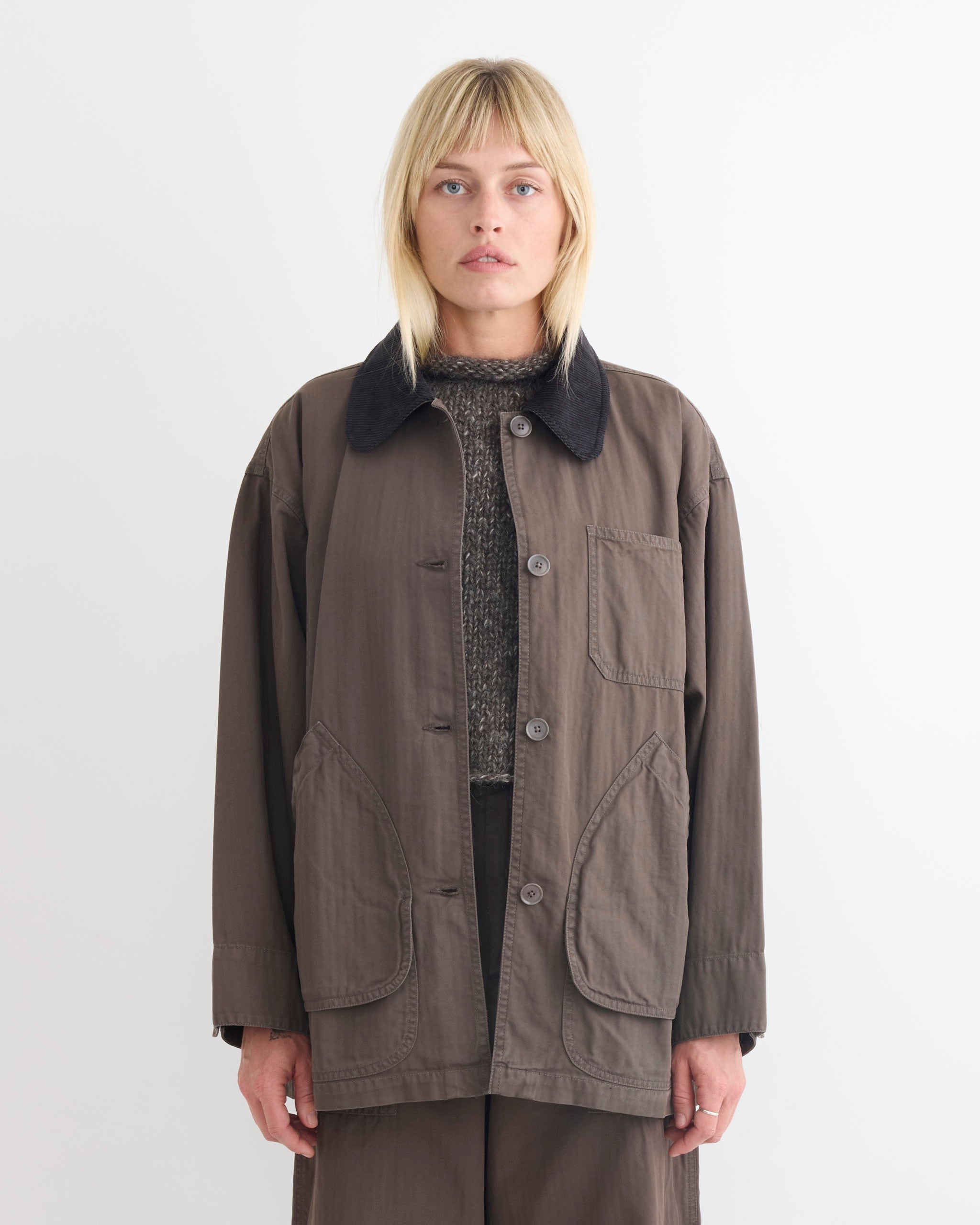 A person with straight blonde hair and bangs wears the Shaina Mote Painter Coat in Ermine, featuring a corduroy collar and large front pockets, over a textured cotton herringbone sweater, against a plain white background.