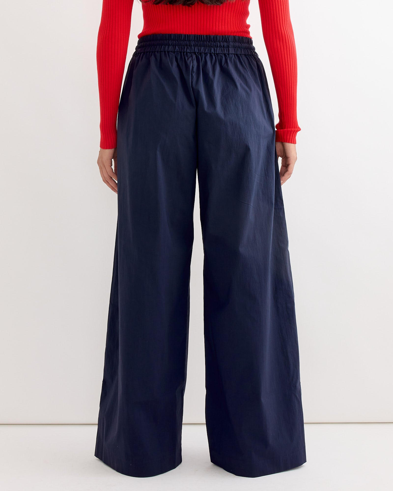 A person wearing a red long-sleeve top and ST. AGNI's Everyday Drawstring Pant in Dark Ink, made from cotton recycled nylon, stands facing away from the camera against a plain background.